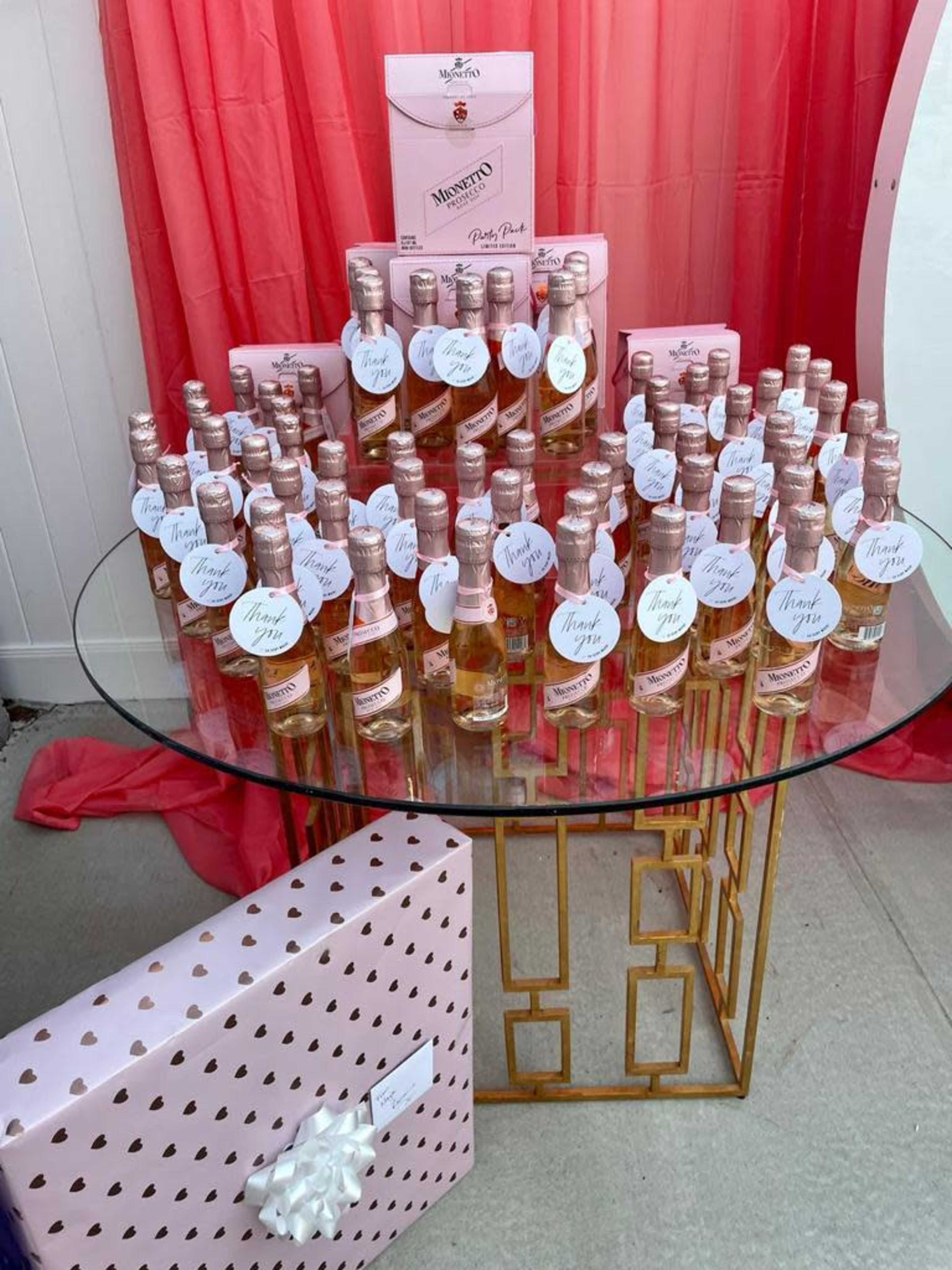A circular table displays an arrangement of bottles of pink wine surrounded by a pink gift box with heart patterns.