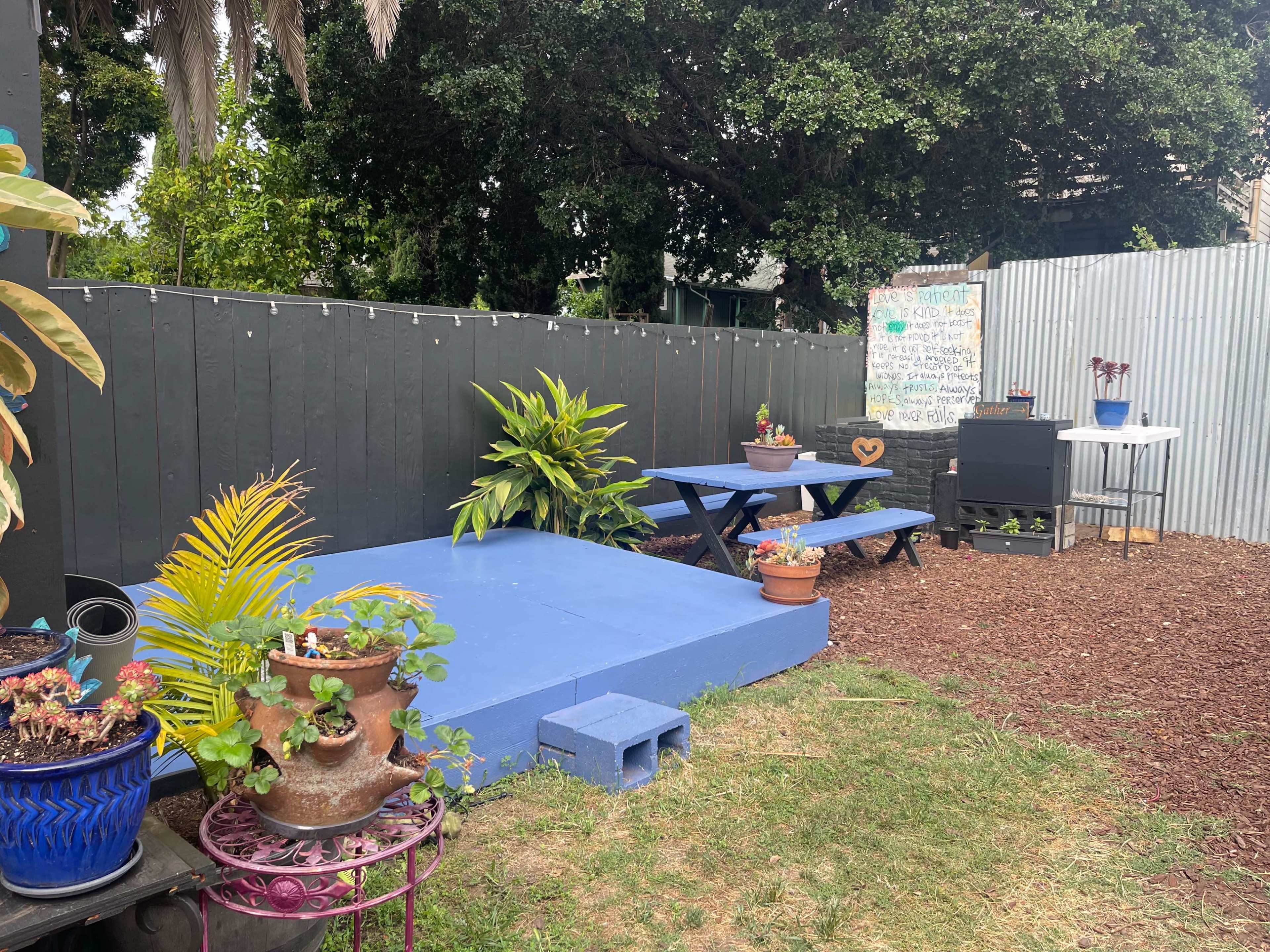 A backyard area featuring a blue platform, a picnic table, potted plants, and a barbecue grill beside a fence.