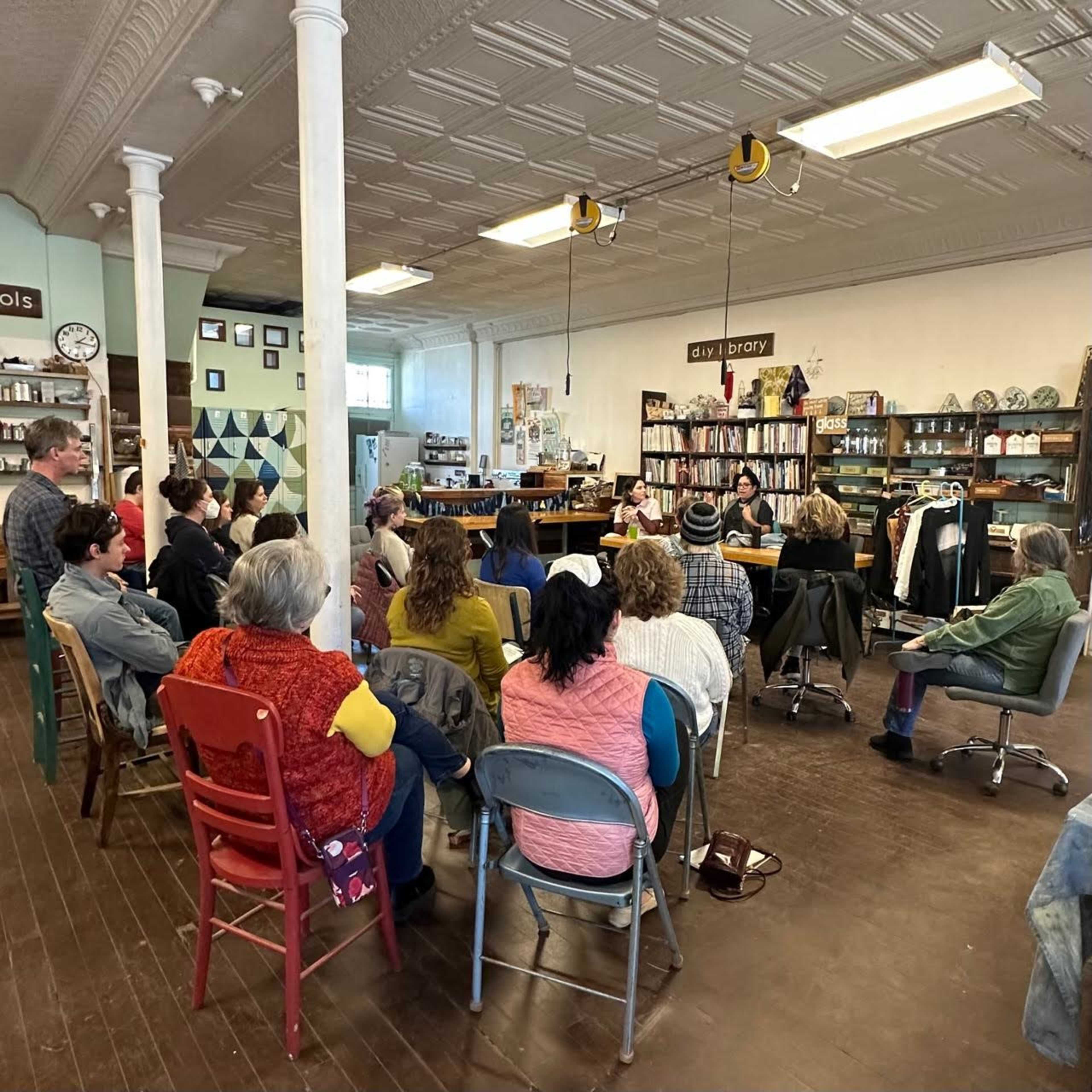 A group of people sits in chairs, facing a table with speakers, in a spacious, well-lit community room filled with bookshelves and decor.