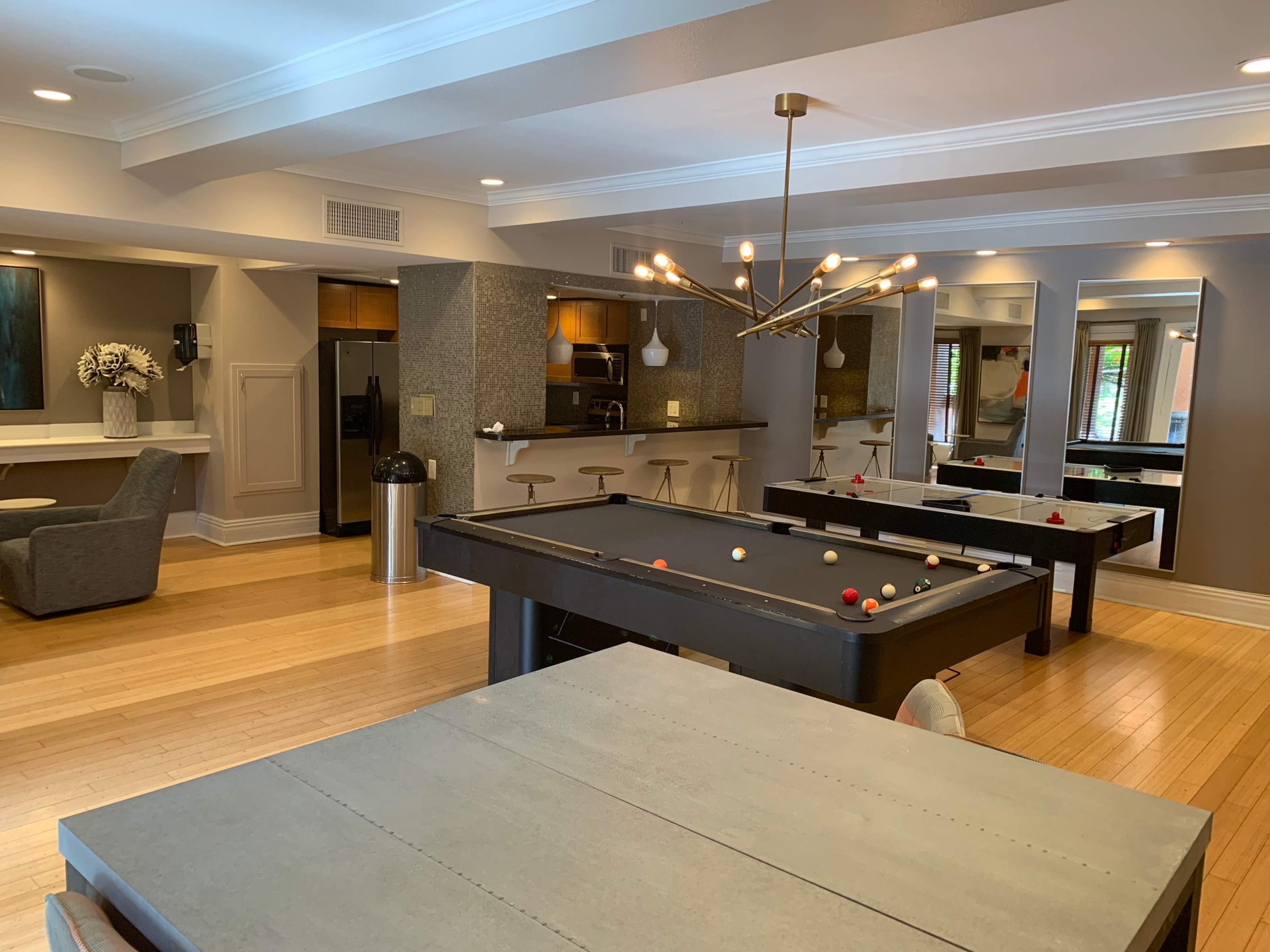 A modern recreational room with a pool table, dining tables, and a kitchen area, featuring hardwood floors and contemporary lighting.