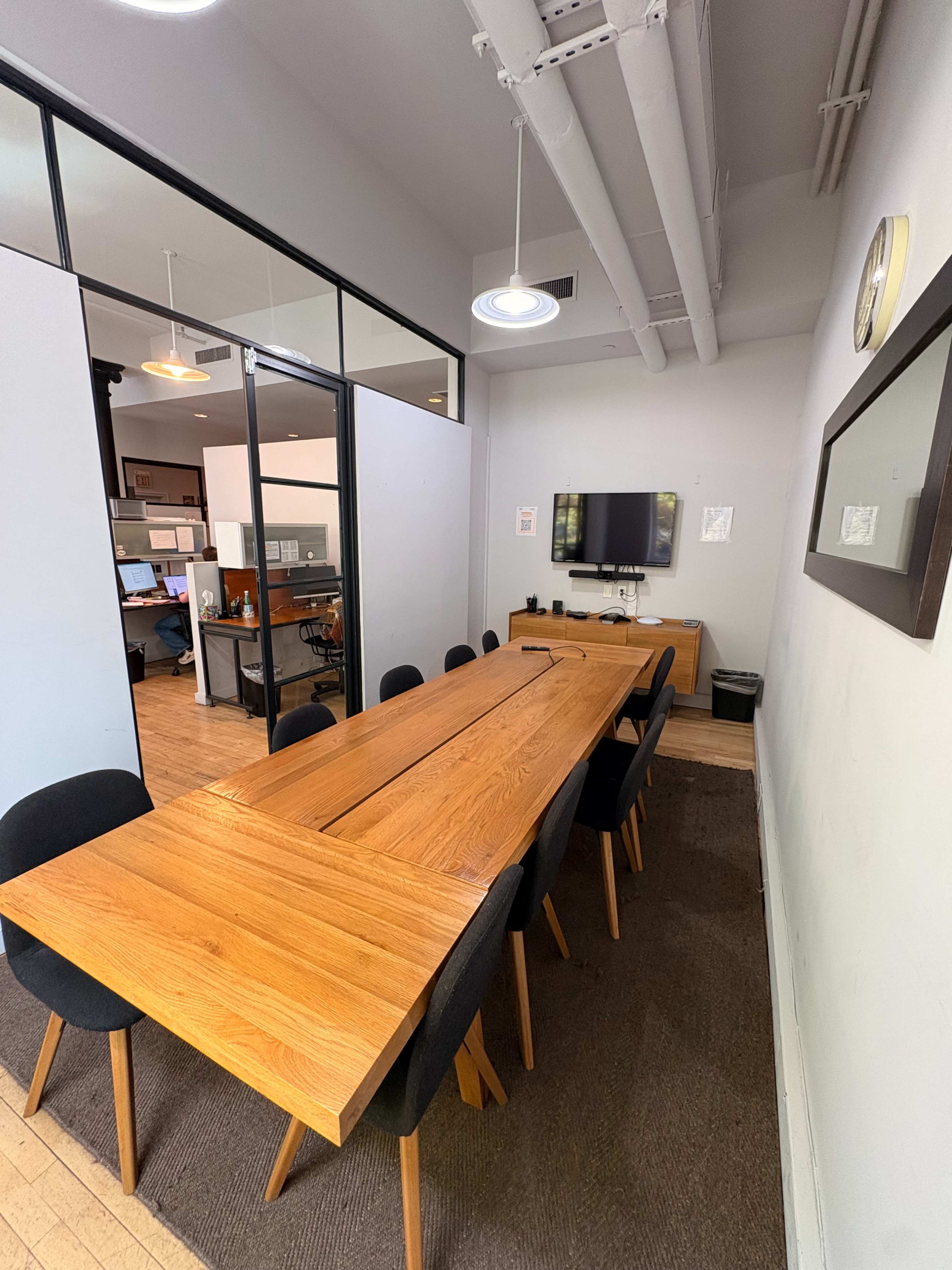 The image shows a modern conference room with a long wooden table surrounded by black chairs, and a television mounted on the wall.