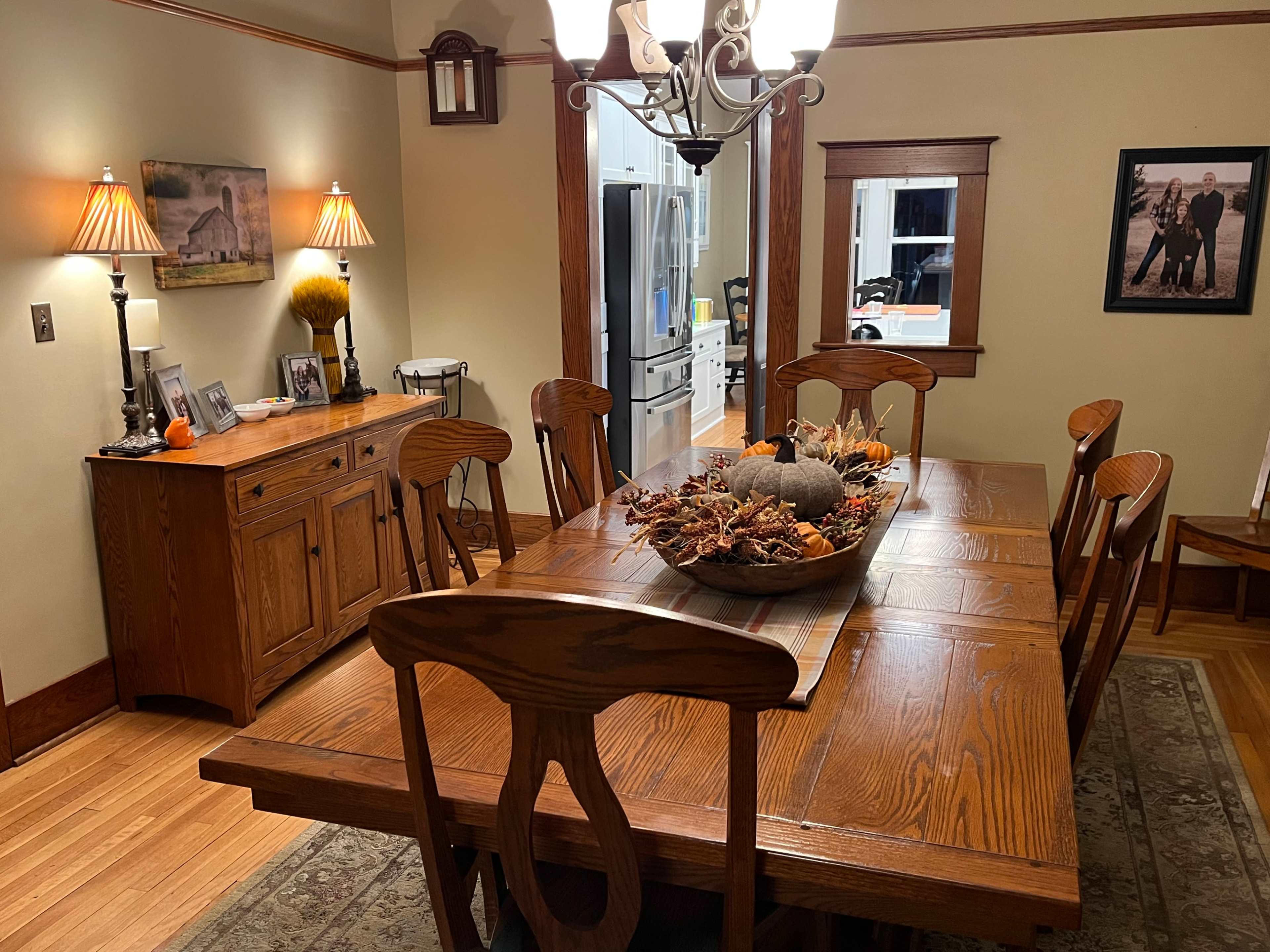 A wooden dining table is set with a decorative centerpiece in an elegantly furnished dining room featuring a sideboard and wall decorations.