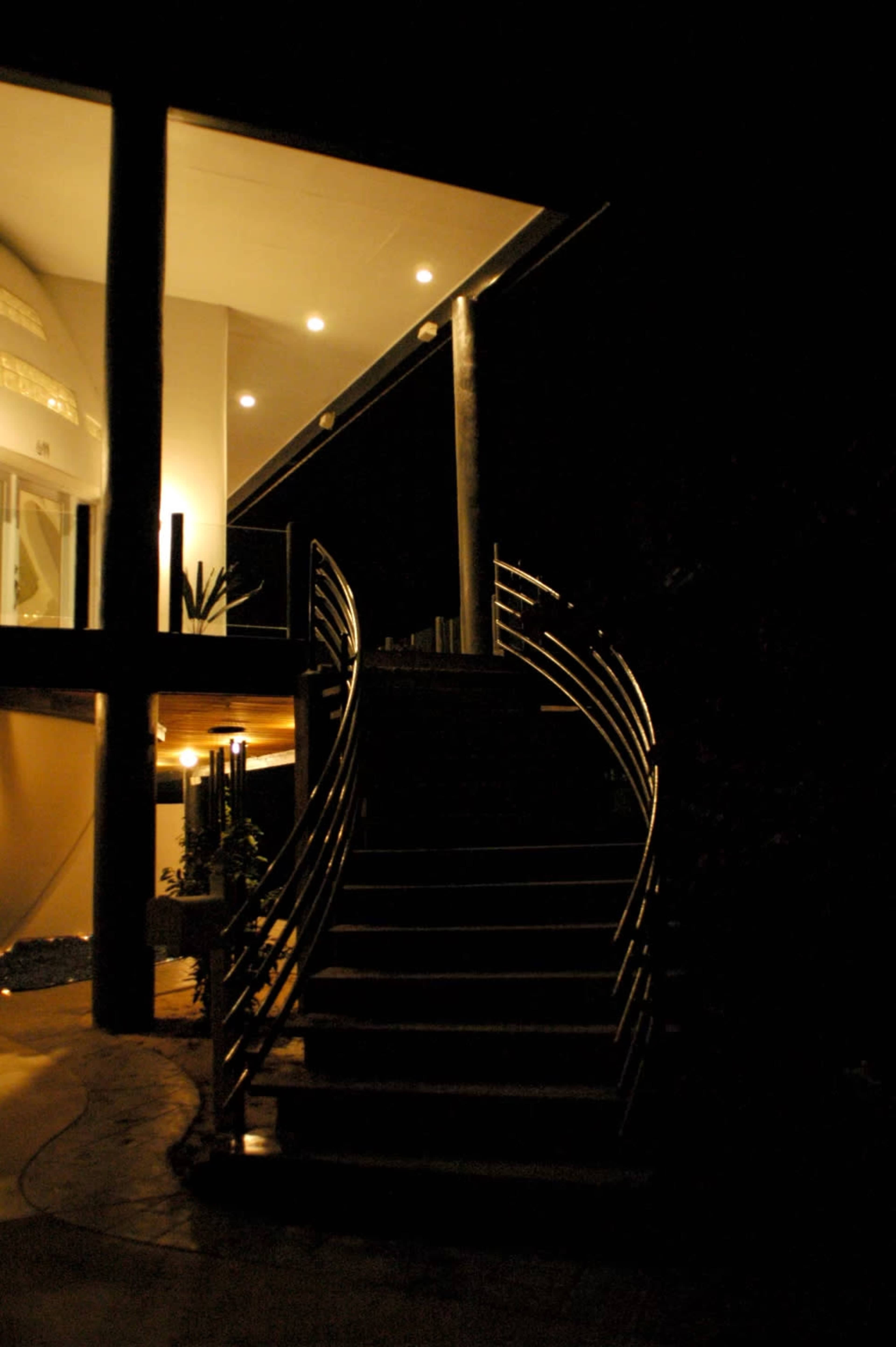 A set of illuminated stairs leads up to a modern house at night, with a sleek railing and surrounding greenery.