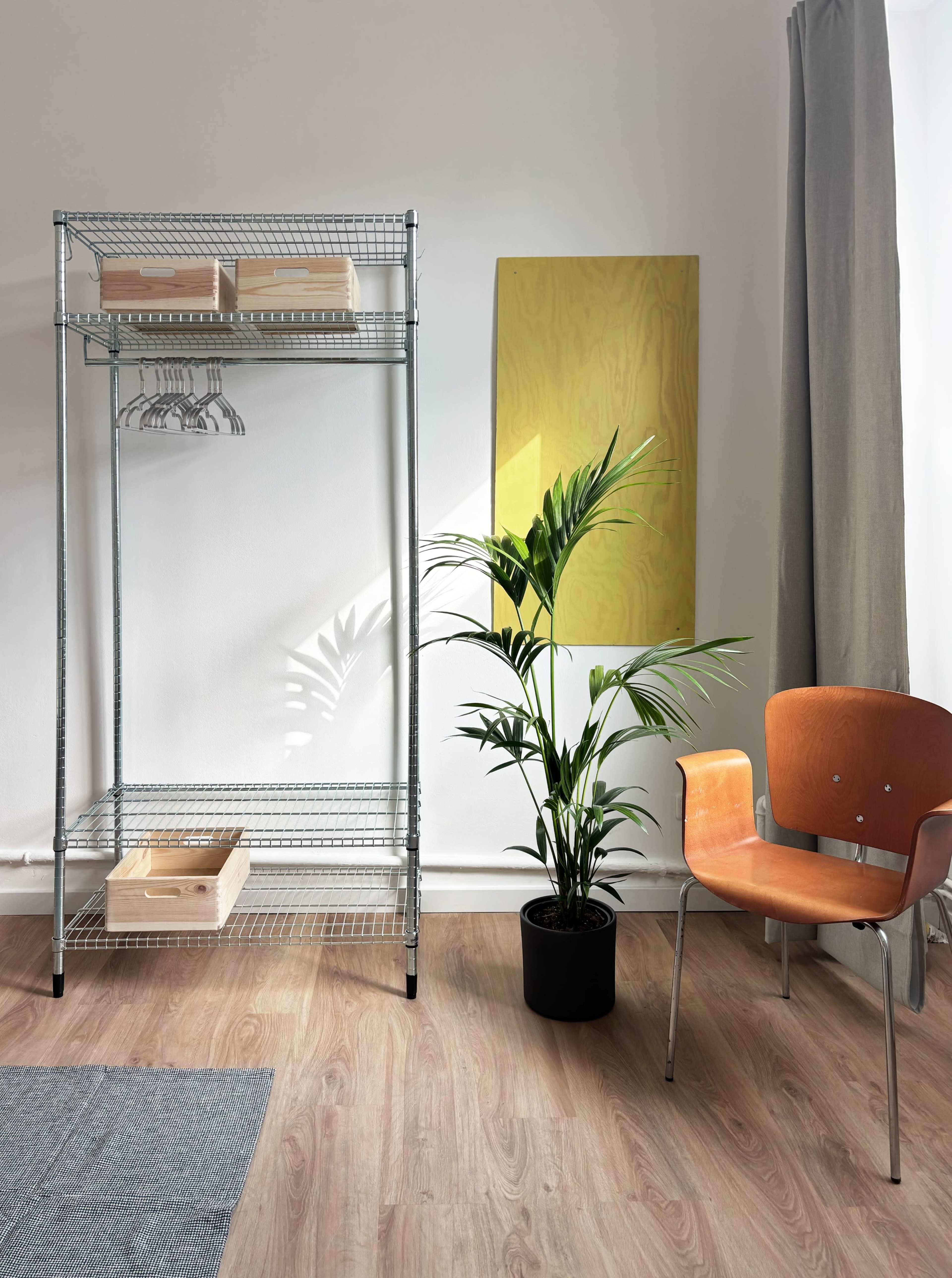 A metal shelving unit with wooden boxes stands beside a potted plant and an orange chair in a minimalist room with wooden flooring.