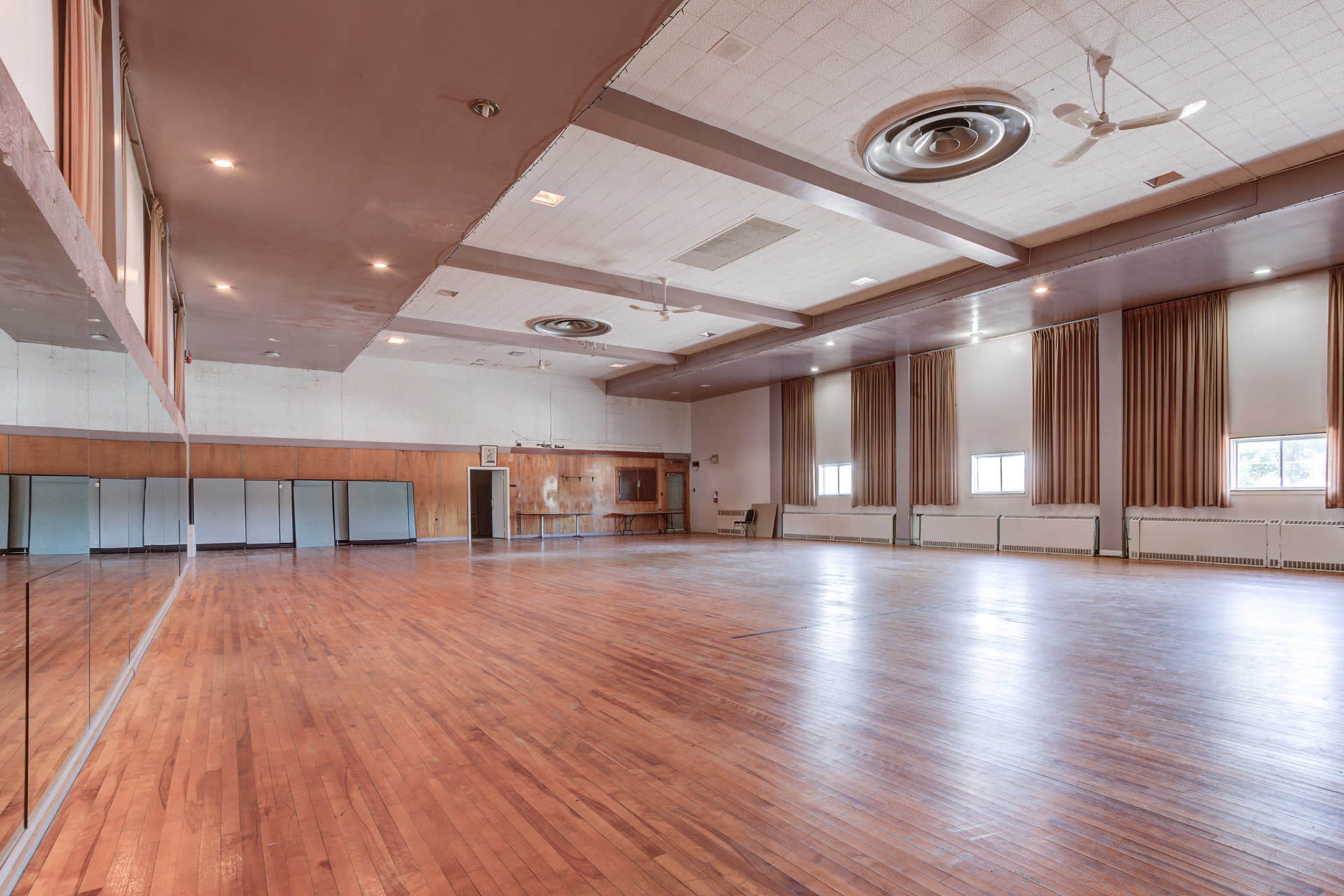 A spacious, empty hall with a polished wooden floor, large windows, and ceiling fans.