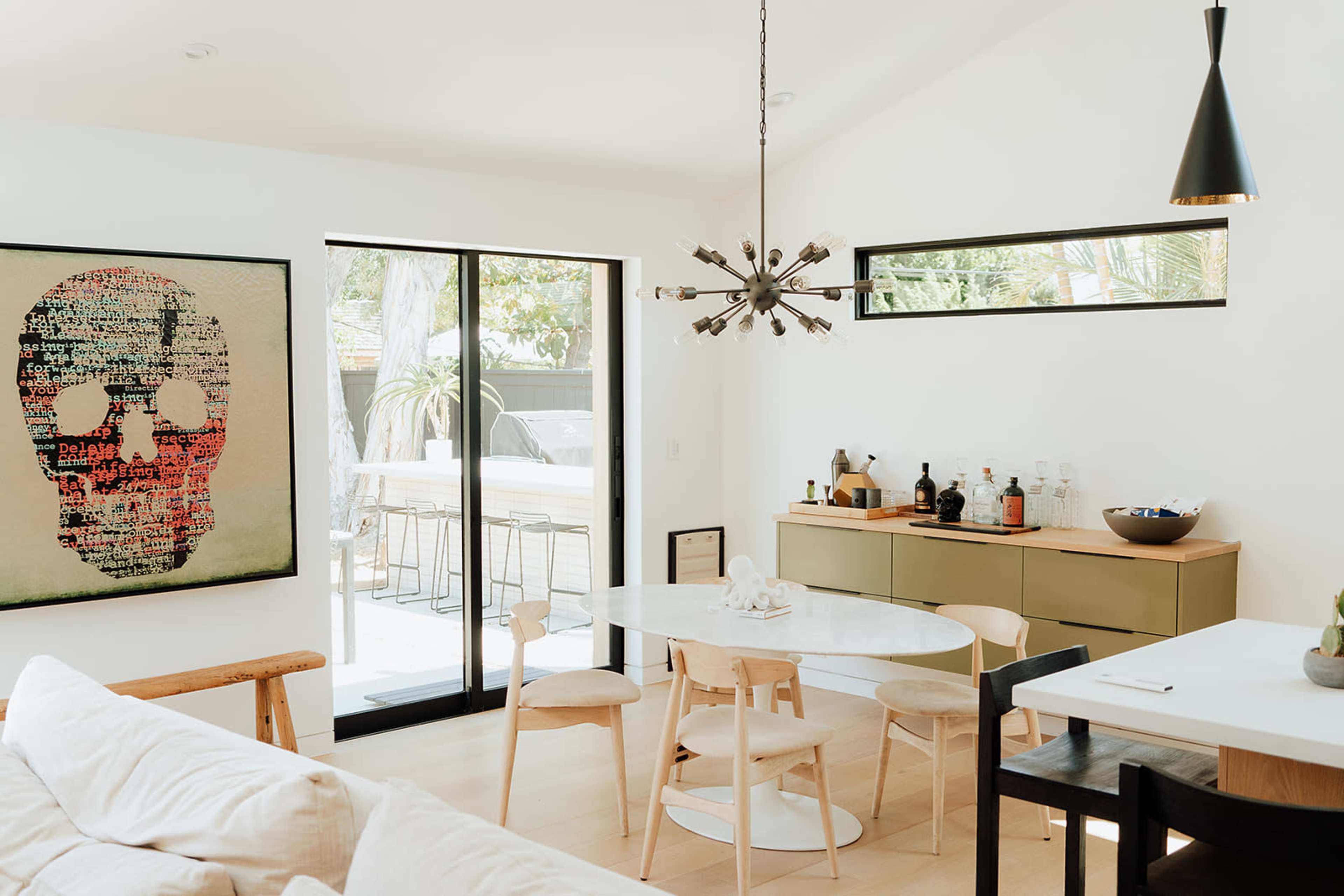 The image shows a modern interior featuring a dining area with a circular marble table surrounded by wooden and black chairs, a sideboard against the wall with various bottles, and a large art piece depicting a skull.