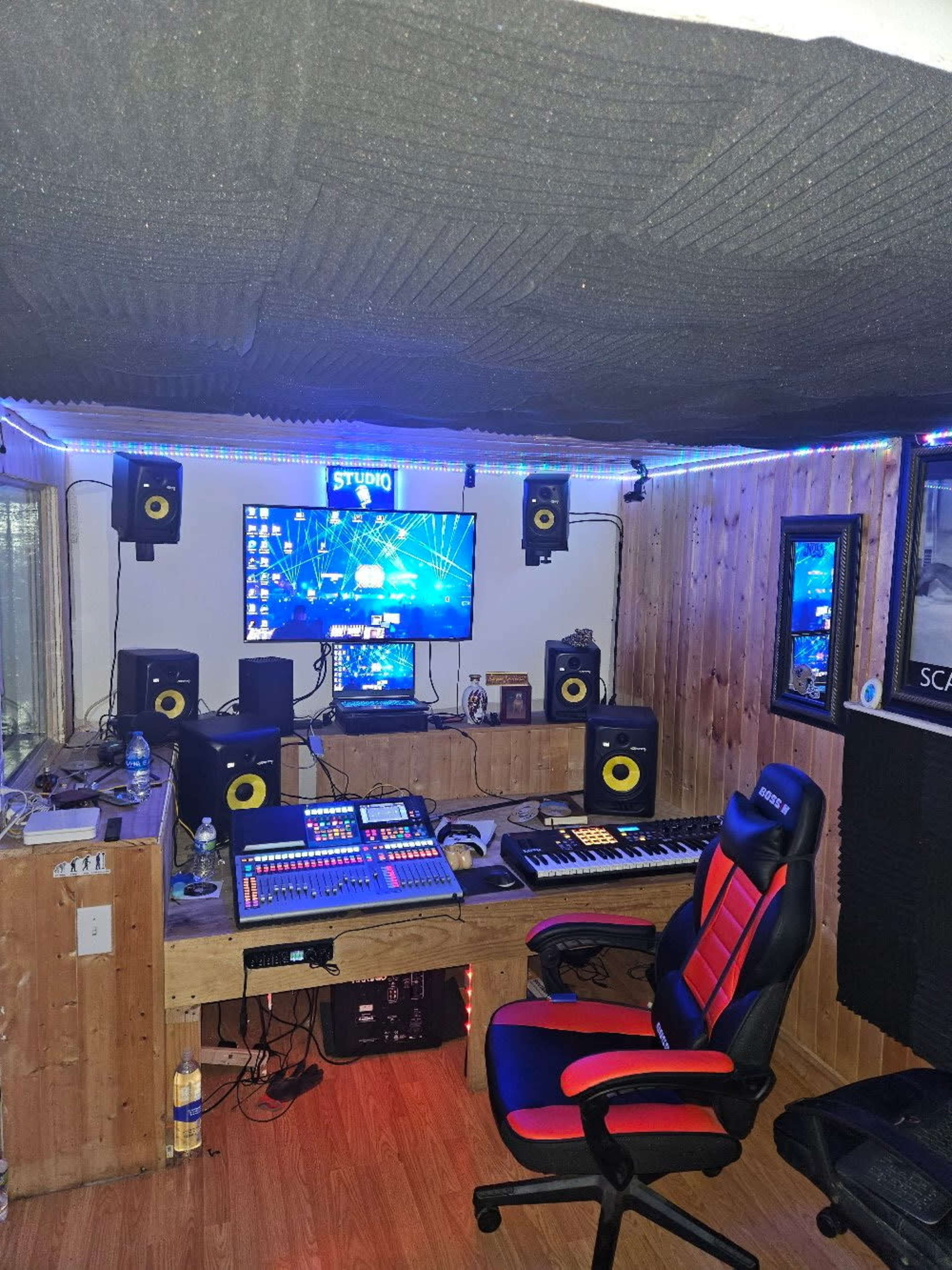 The image shows a music production studio featuring a mixing console, computer monitor, speakers, keyboard, and soundproofed walls.