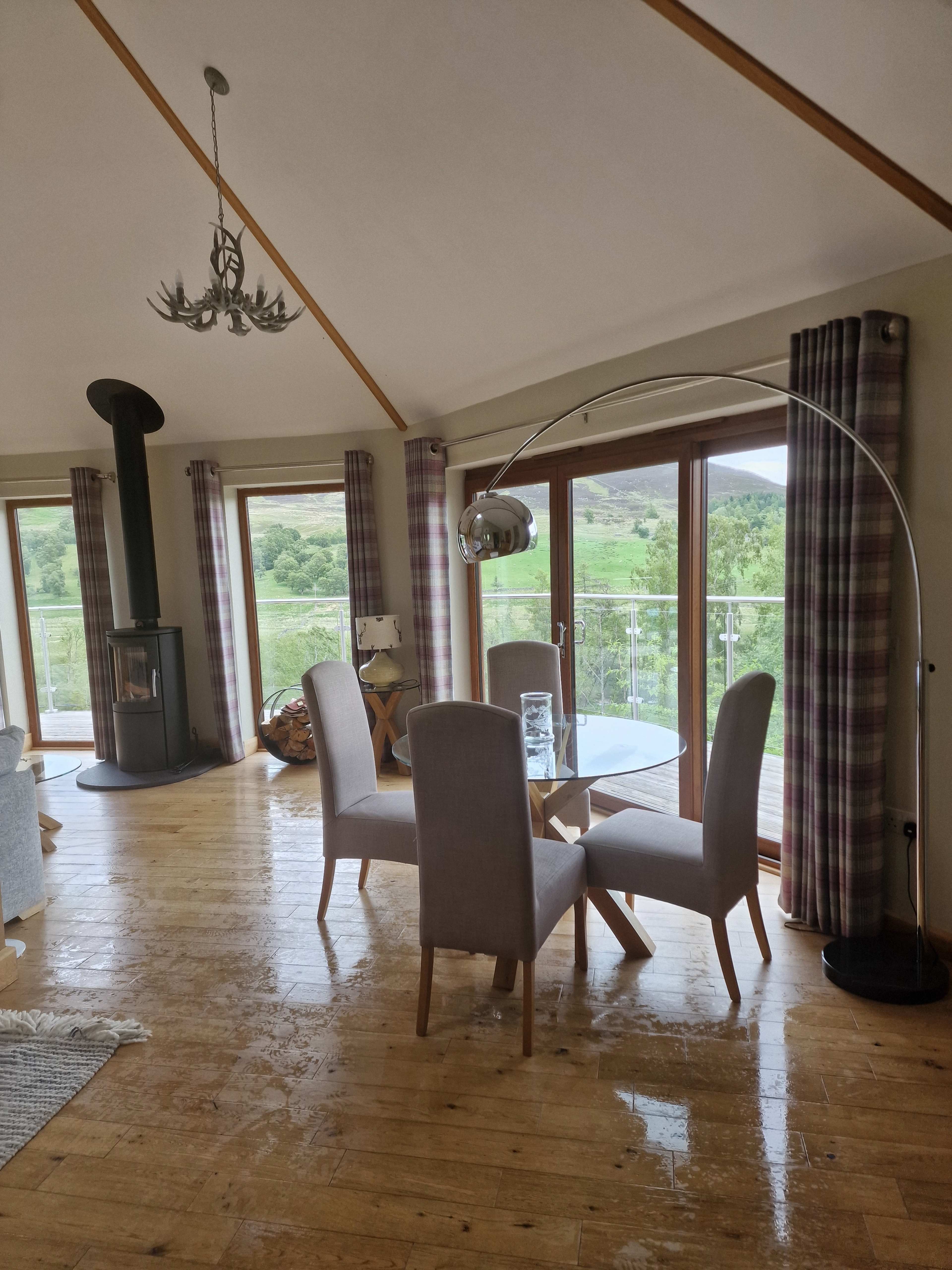 A dining area features a round table surrounded by four upholstered chairs, with large windows showcasing a green landscape.