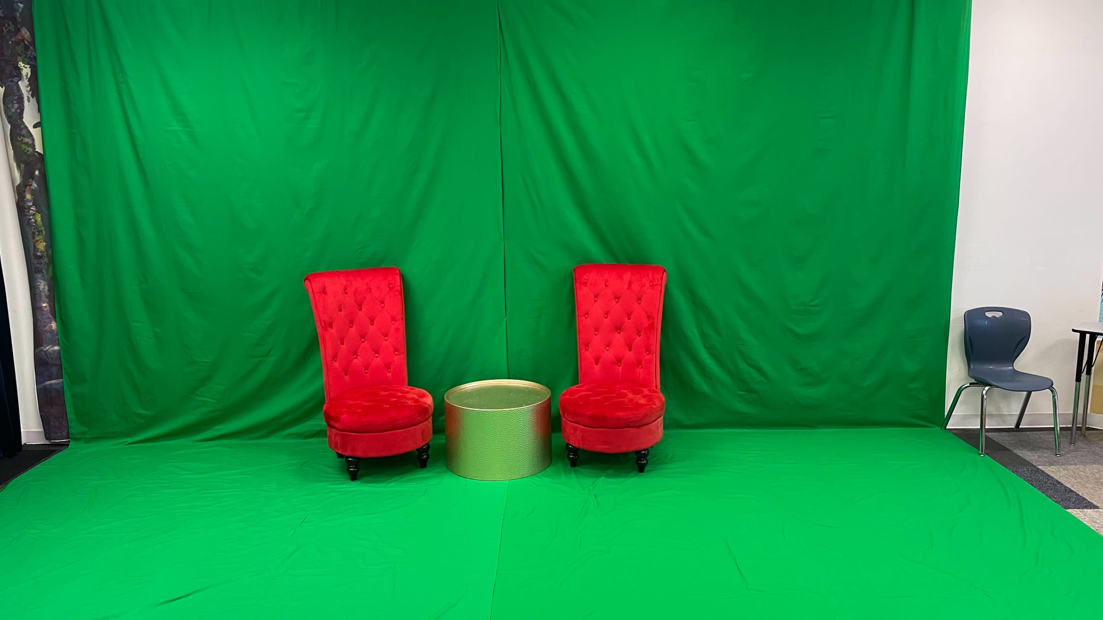 A green screen setup featuring two red velvet chairs and a small gold table positioned in front of a green backdrop.