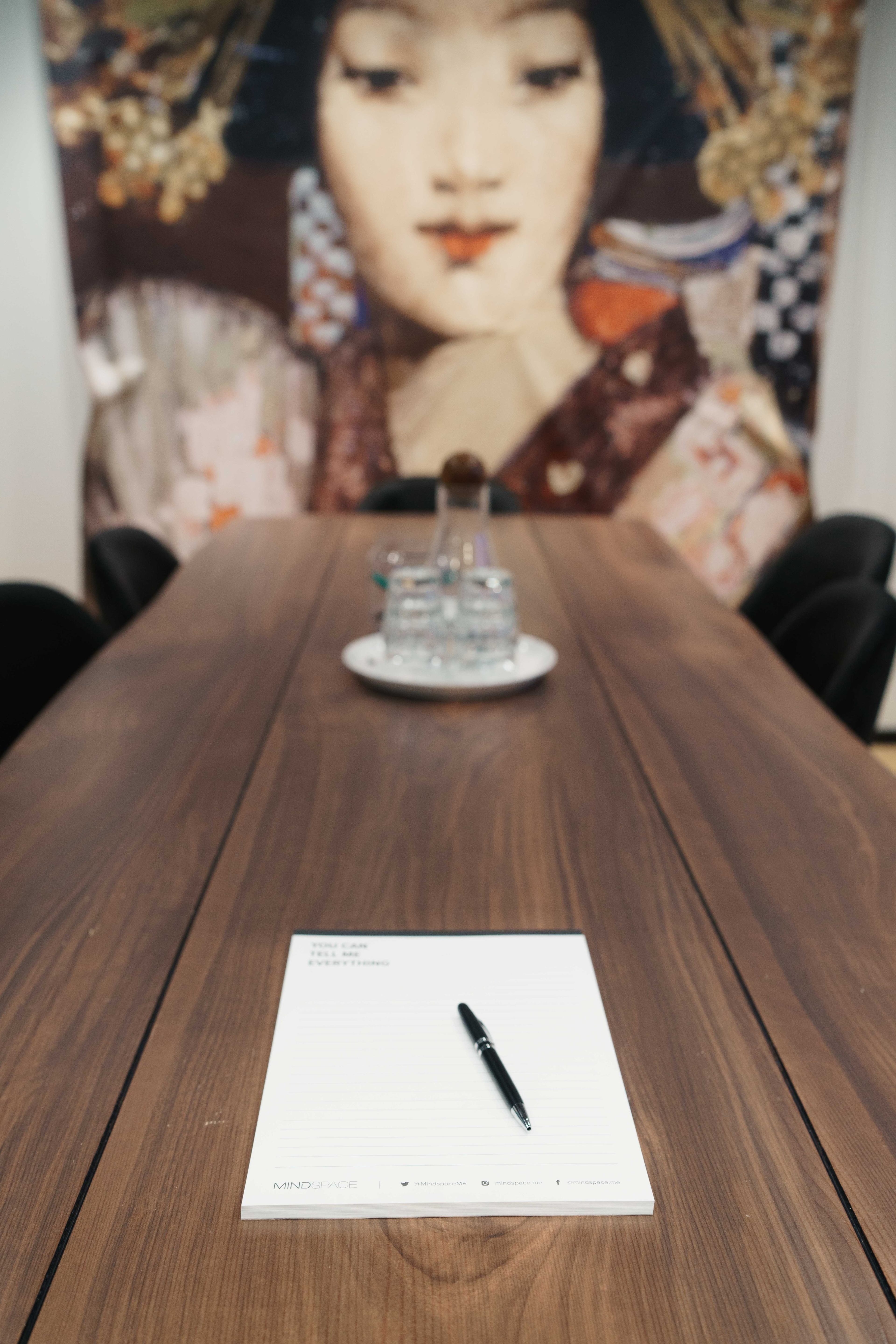 A large wooden table with a notepad and pen is positioned in front of a colorful wall art featuring a woman's face.