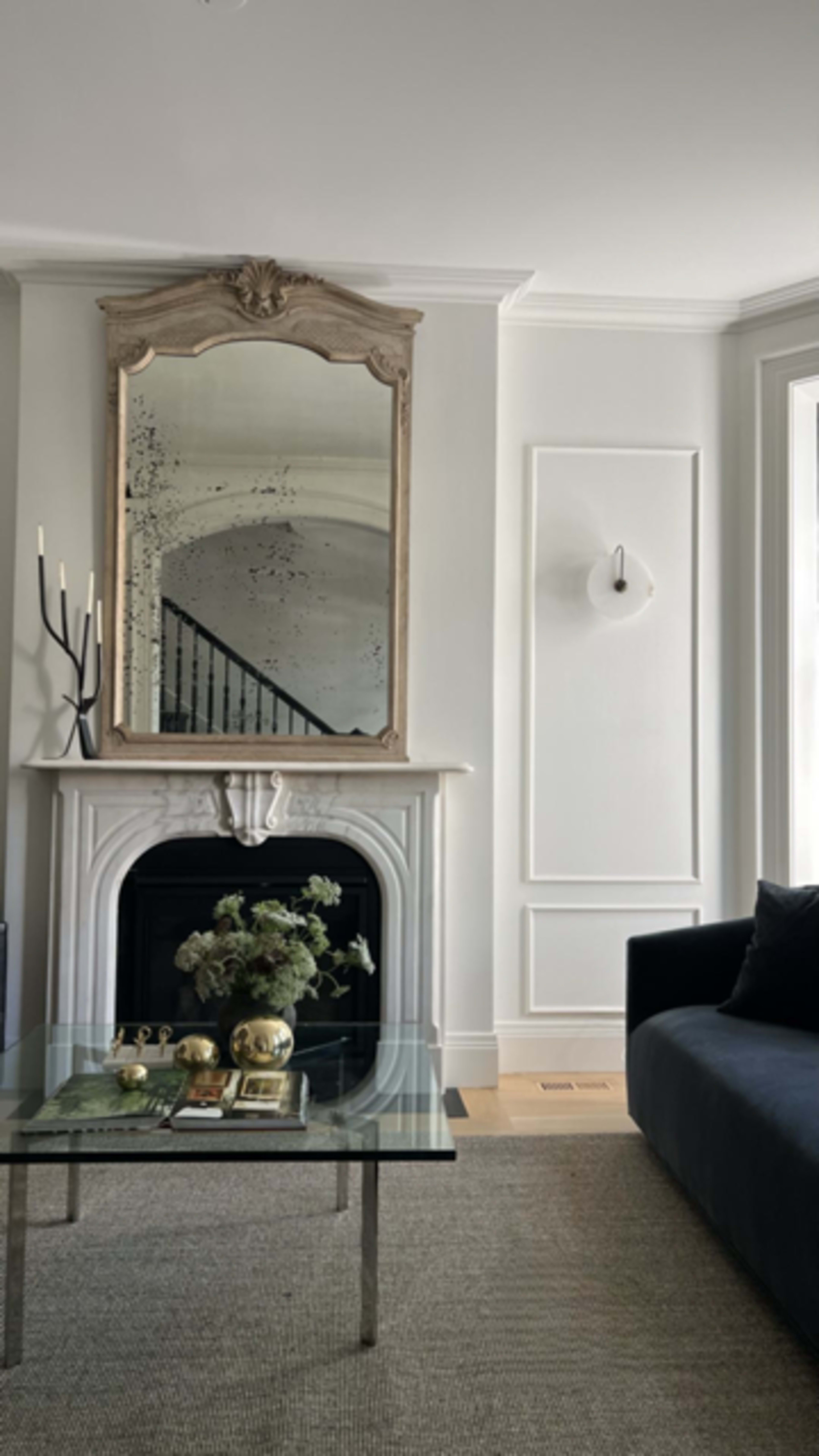 A modern living room featuring a glass coffee table in front of a black sofa, a decorative mirror above a white mantel, and a staircase visible in the background.