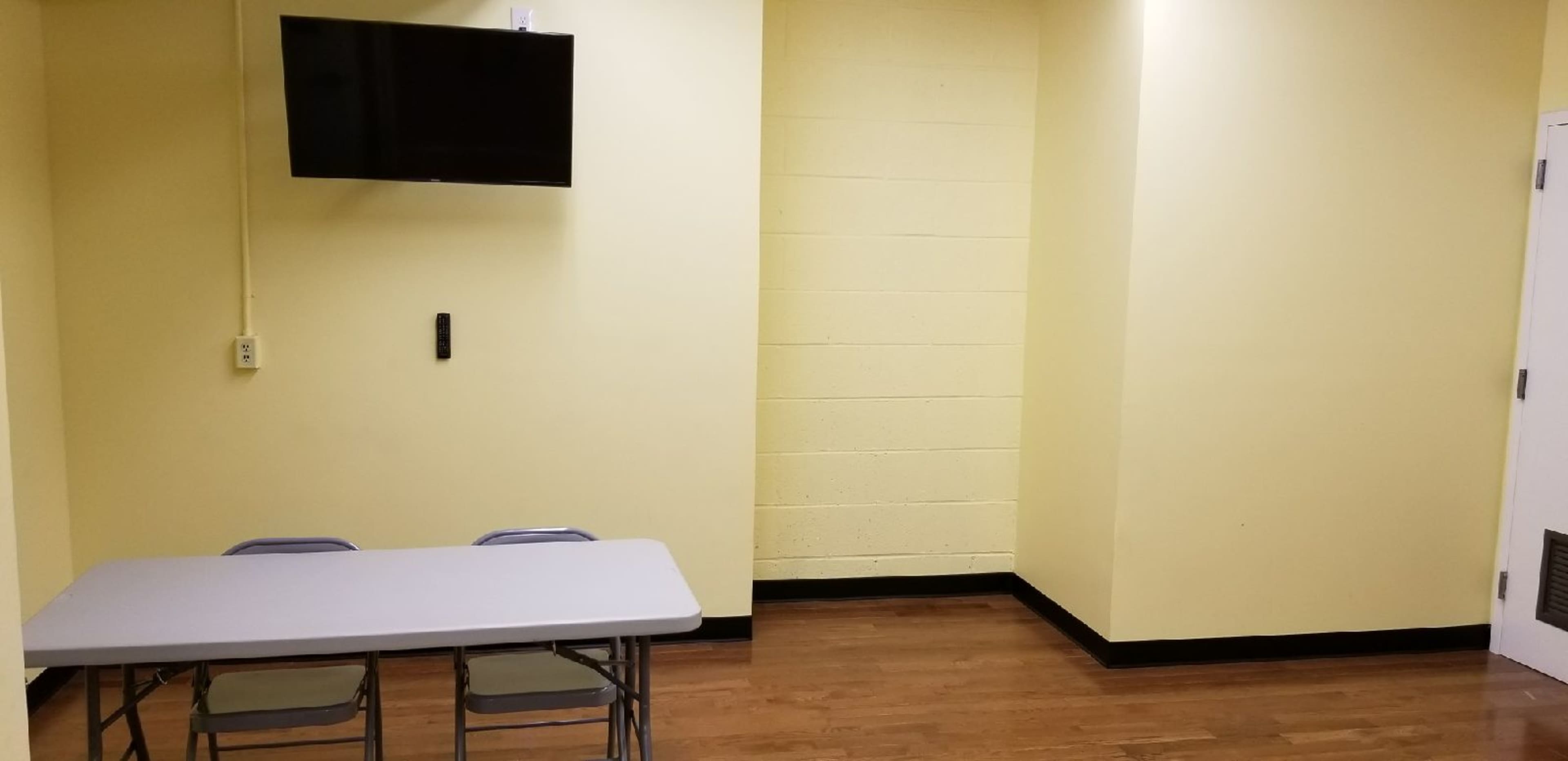 The image shows a small, yellow-walled room with a table and chairs, a mounted TV, and a door in one corner.