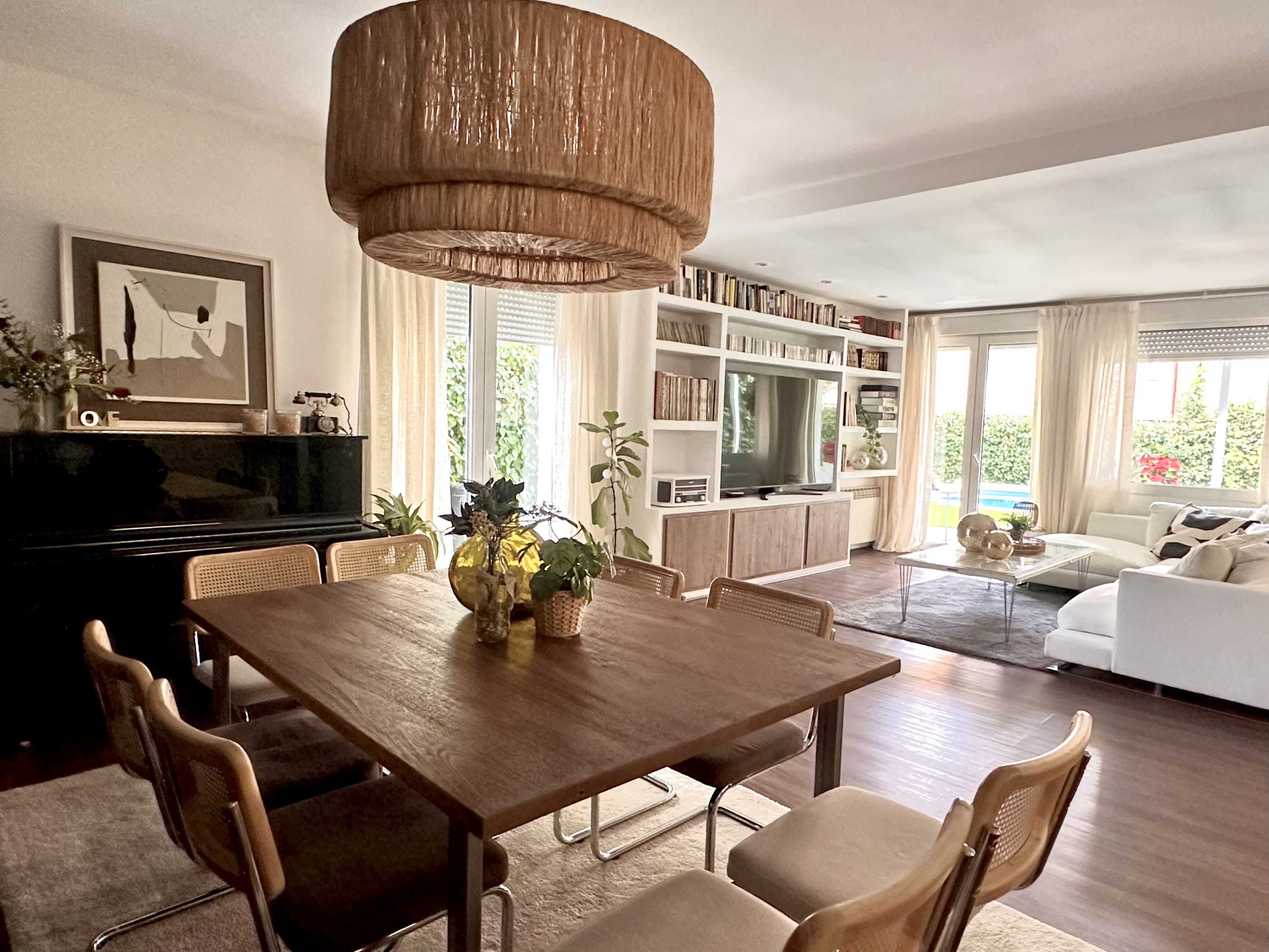 A dining area features a wooden table surrounded by chairs, with a large woven light fixture above and a living room visible in the background.