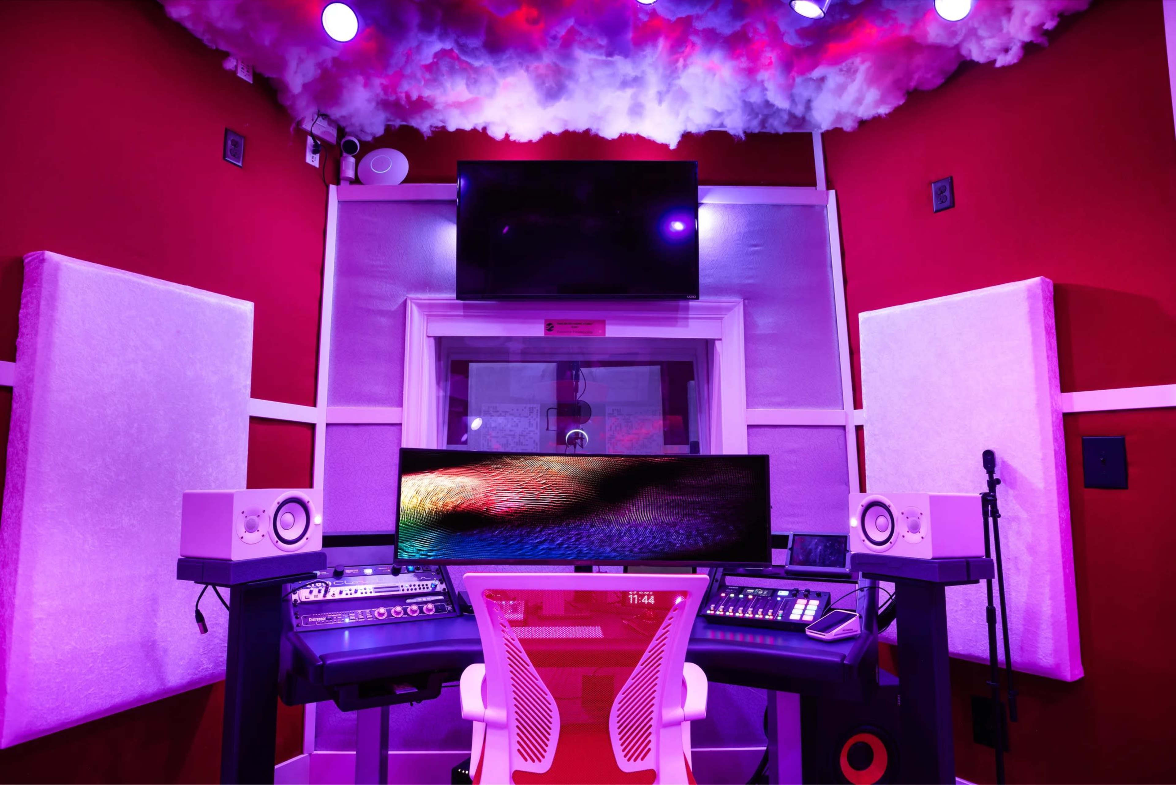 A music studio features a central desk with audio equipment, a large monitor, and soundproofing panels against bright red walls.