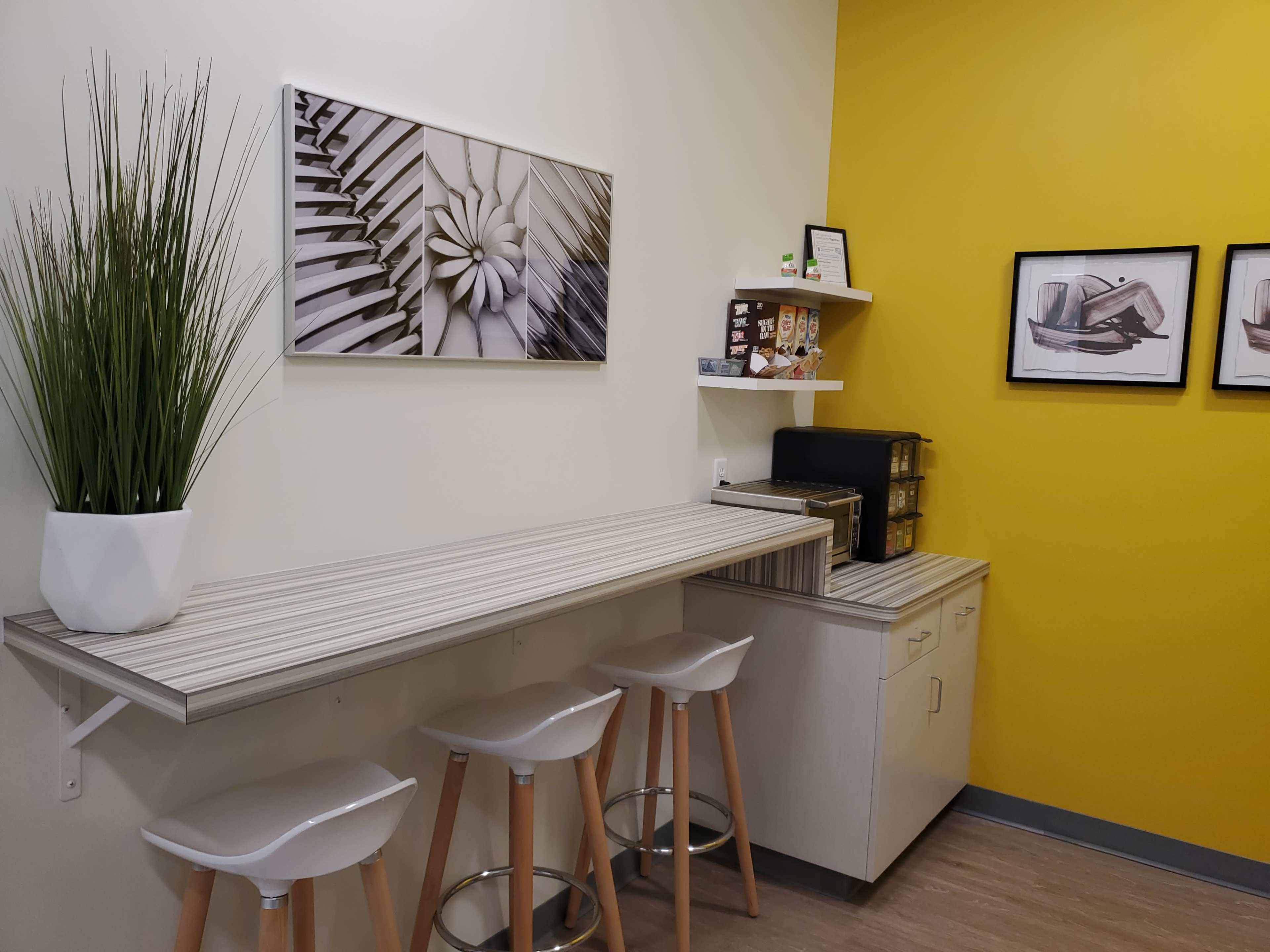 A small kitchenette area with a long counter, three stools, a wall-mounted art piece, and shelves containing snacks and a coffee machine, set against a yellow wall.