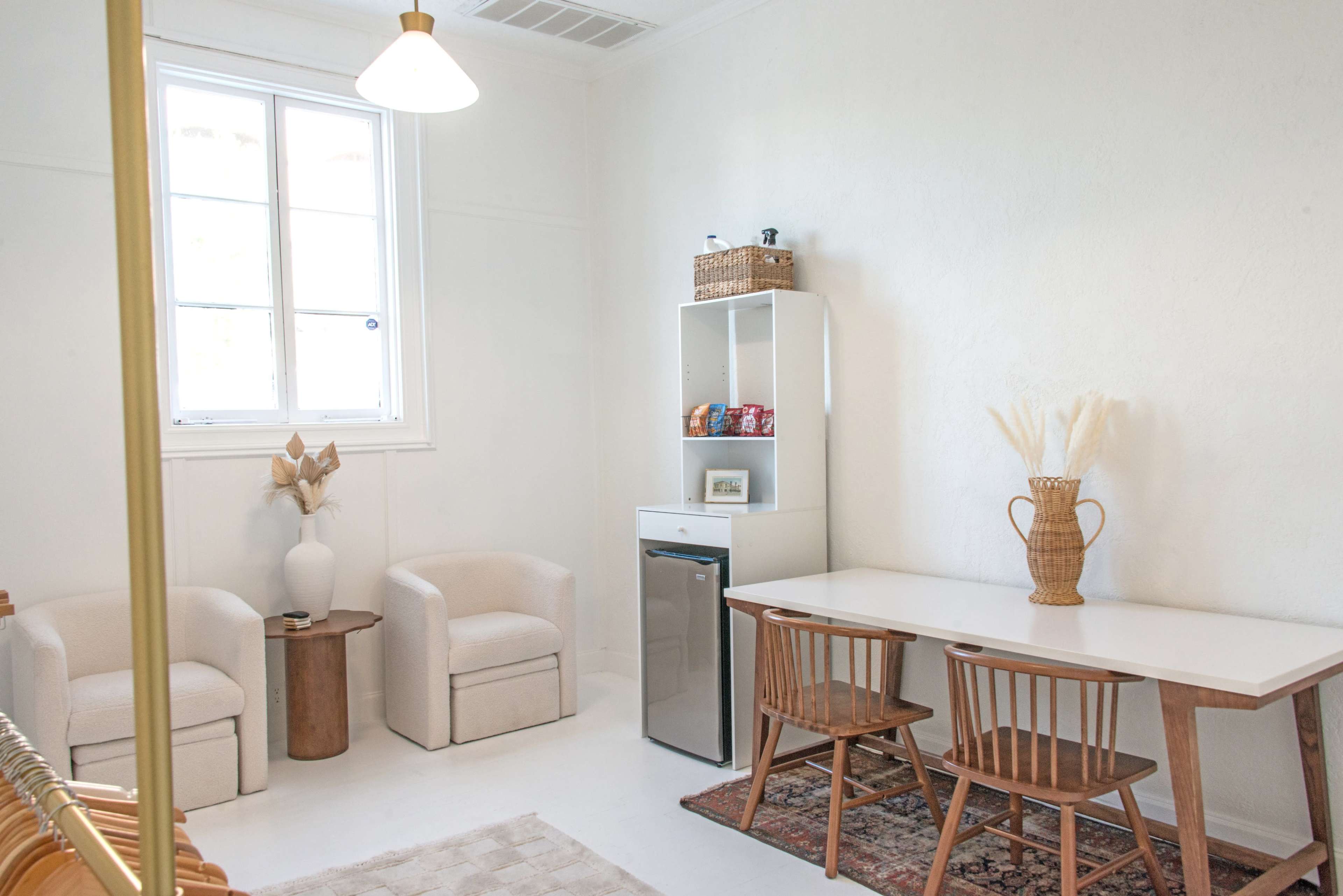 The image shows a bright, minimalist room featuring two upholstered chairs, a small table with two wooden chairs, a small refrigerator, and a shelving unit with decorative items.