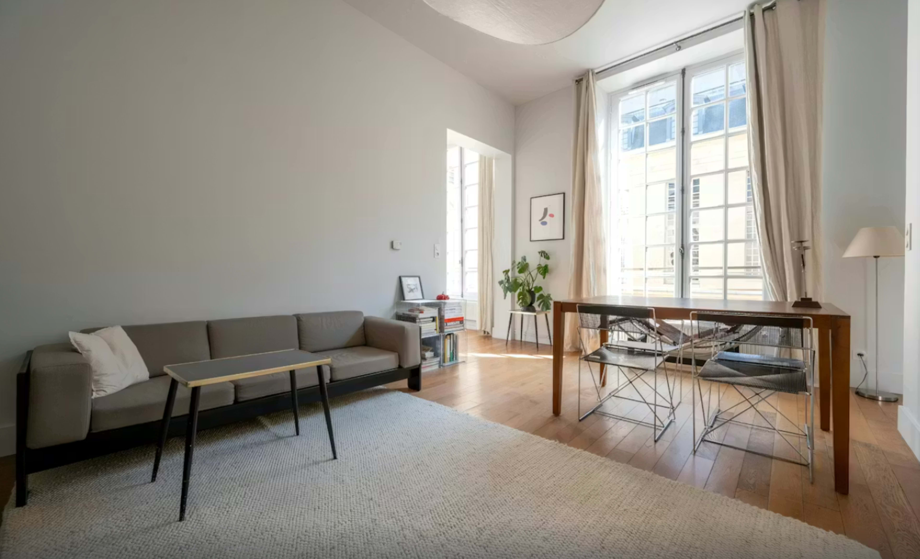 The image shows a minimalistic living space with a grey sofa, a wooden table with metal chairs, and large windows allowing natural light to illuminate the room.