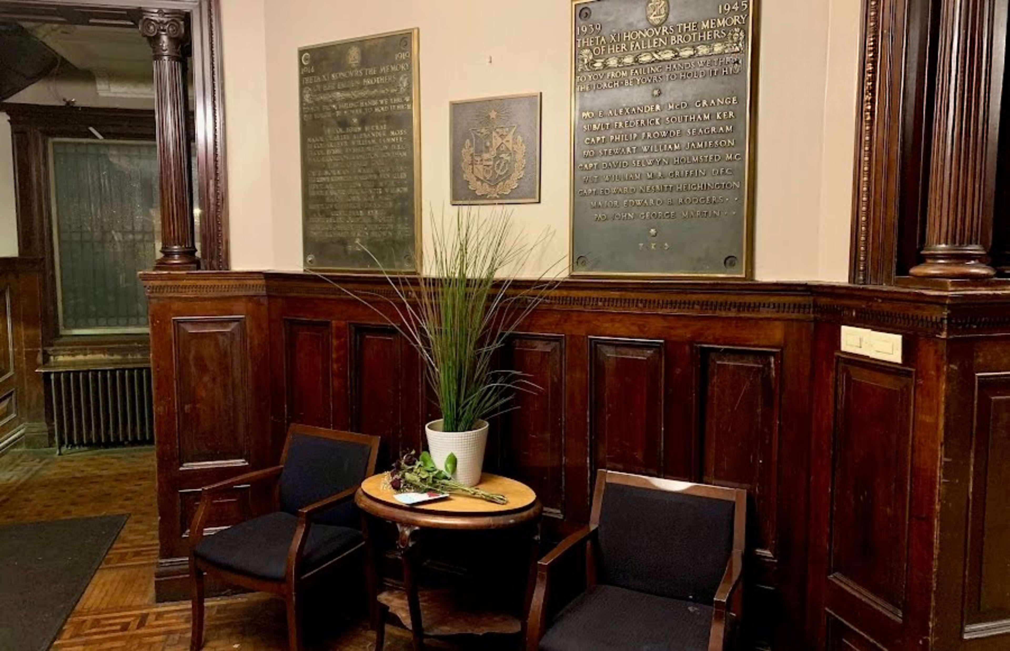 The image shows a corner of a room featuring two wooden chairs and a small round table with a plant on it, surrounded by dark wooden paneling and plaques on the wall.