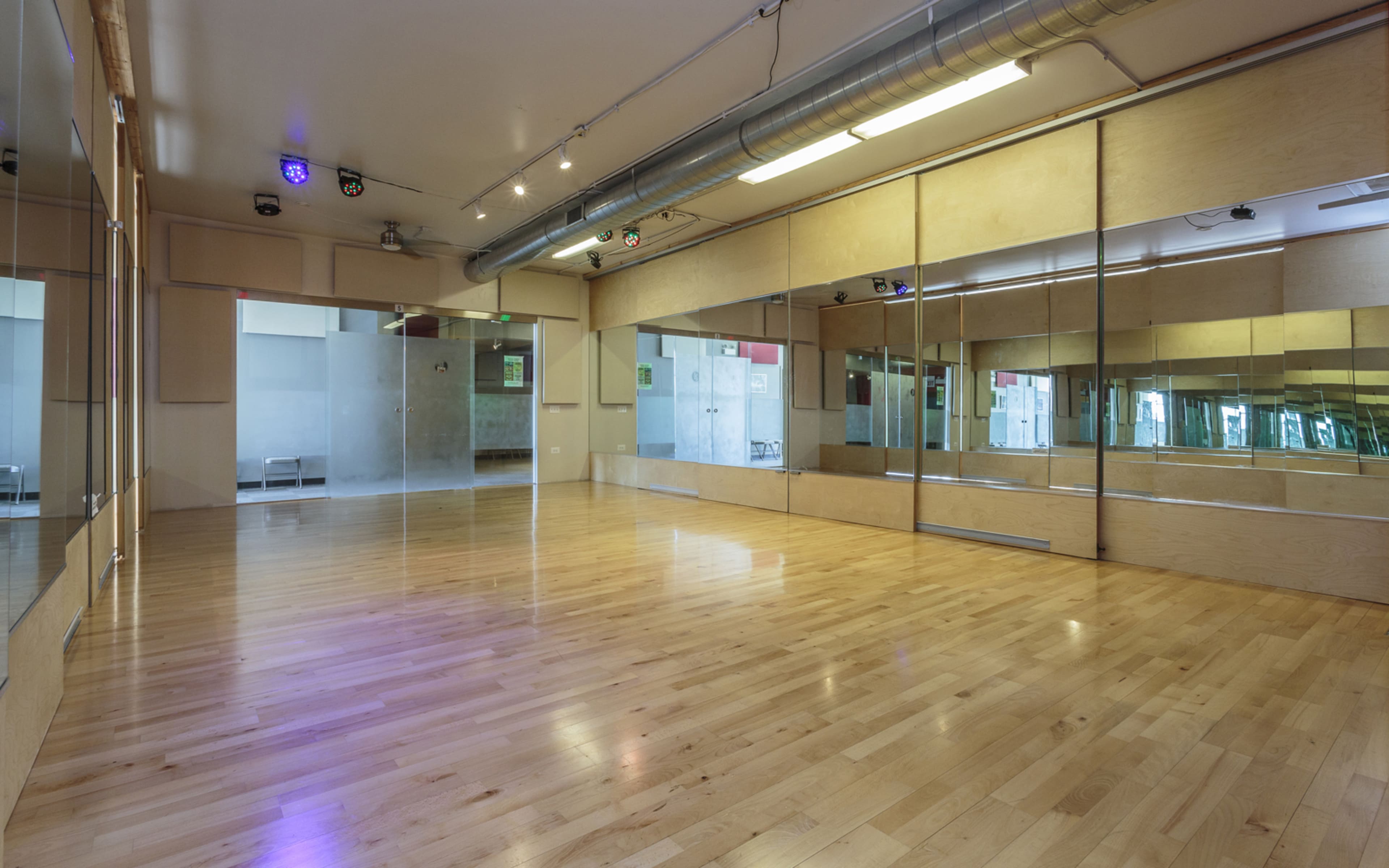A spacious dance studio with wooden flooring, mirrored walls, and overhead lighting.