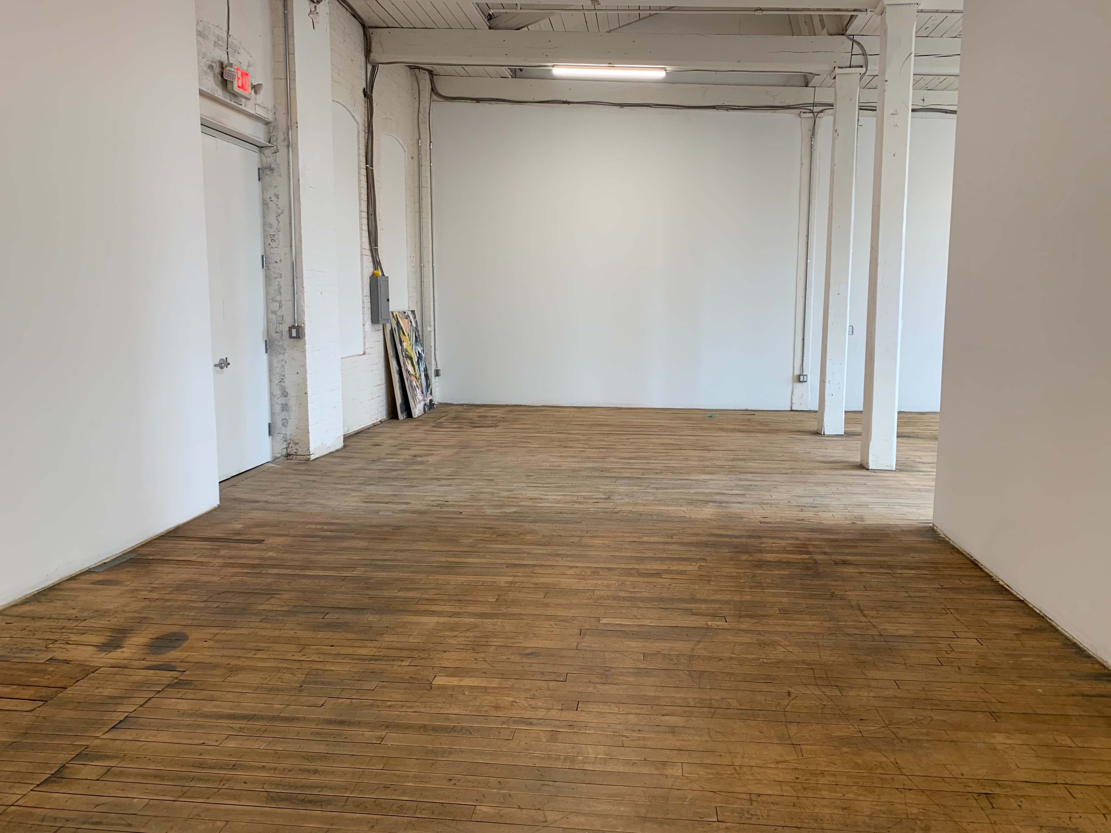 An empty room with wooden floors and white walls, featuring a door on one side and a few framed artworks leaning against the wall.