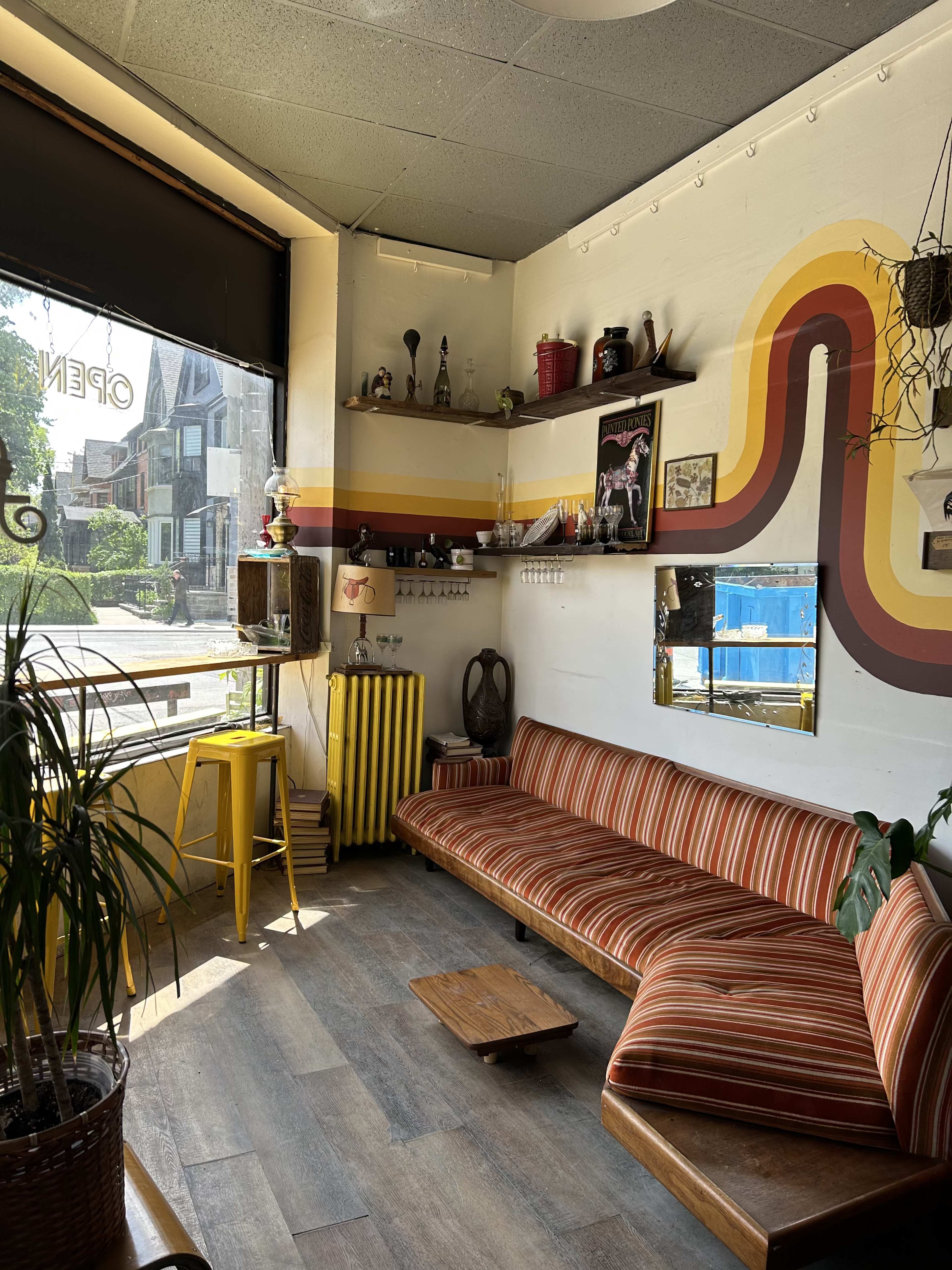 A cozy café interior with a striped sofa, wooden table, potted plants, and vintage decorations against a brightly painted wall, all illuminated by natural light from a large window.