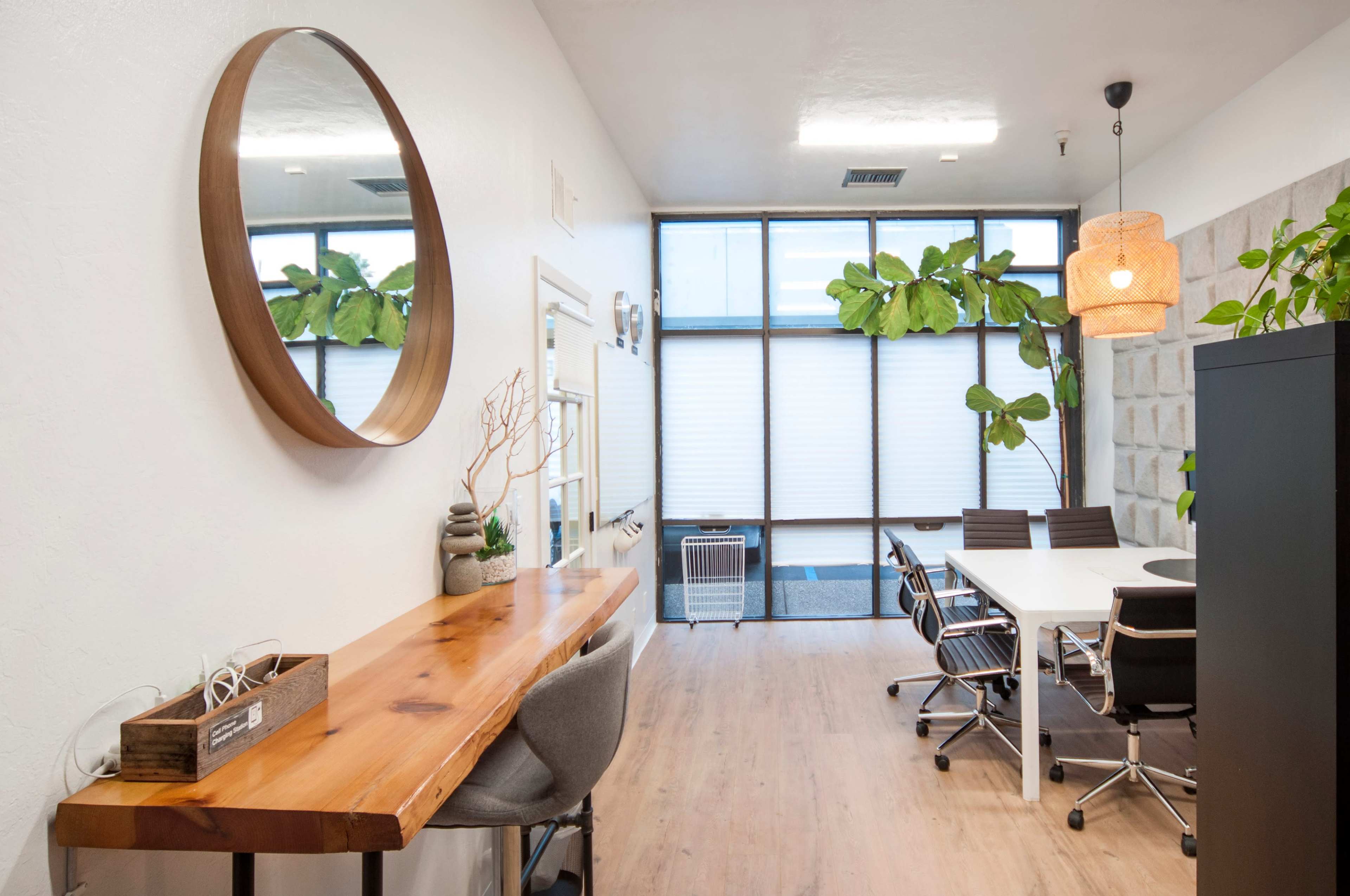A modern office space with a wooden table, a large round mirror, and a meeting area featuring a white table and black chairs, complemented by plants and large windows.
