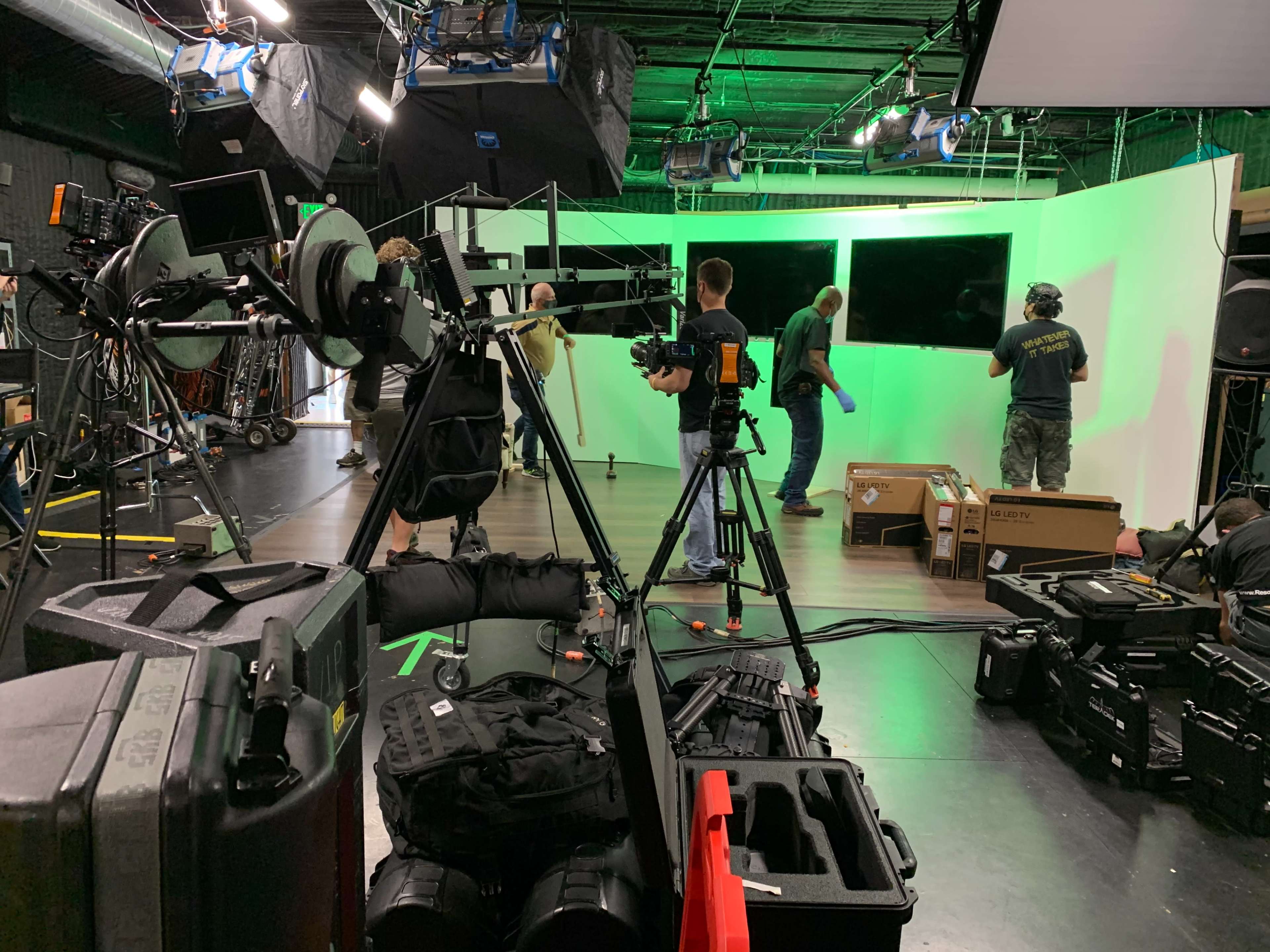 A film crew sets up equipment on a studio set with green screens and various cameras in the background.