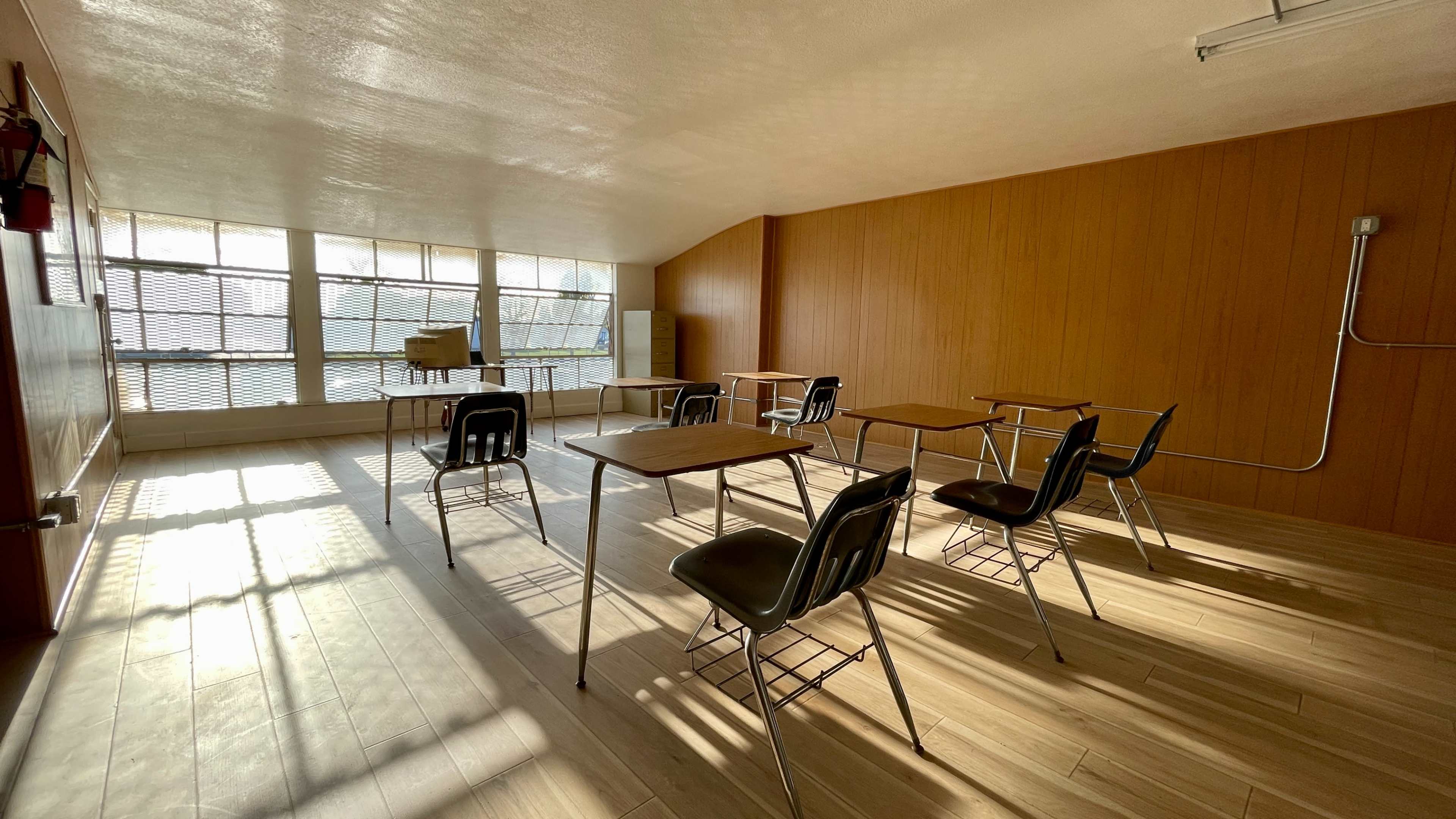 An empty classroom with wooden walls, metal desks, and chairs arranged in a spacious layout, illuminated by sunlight through large windows.