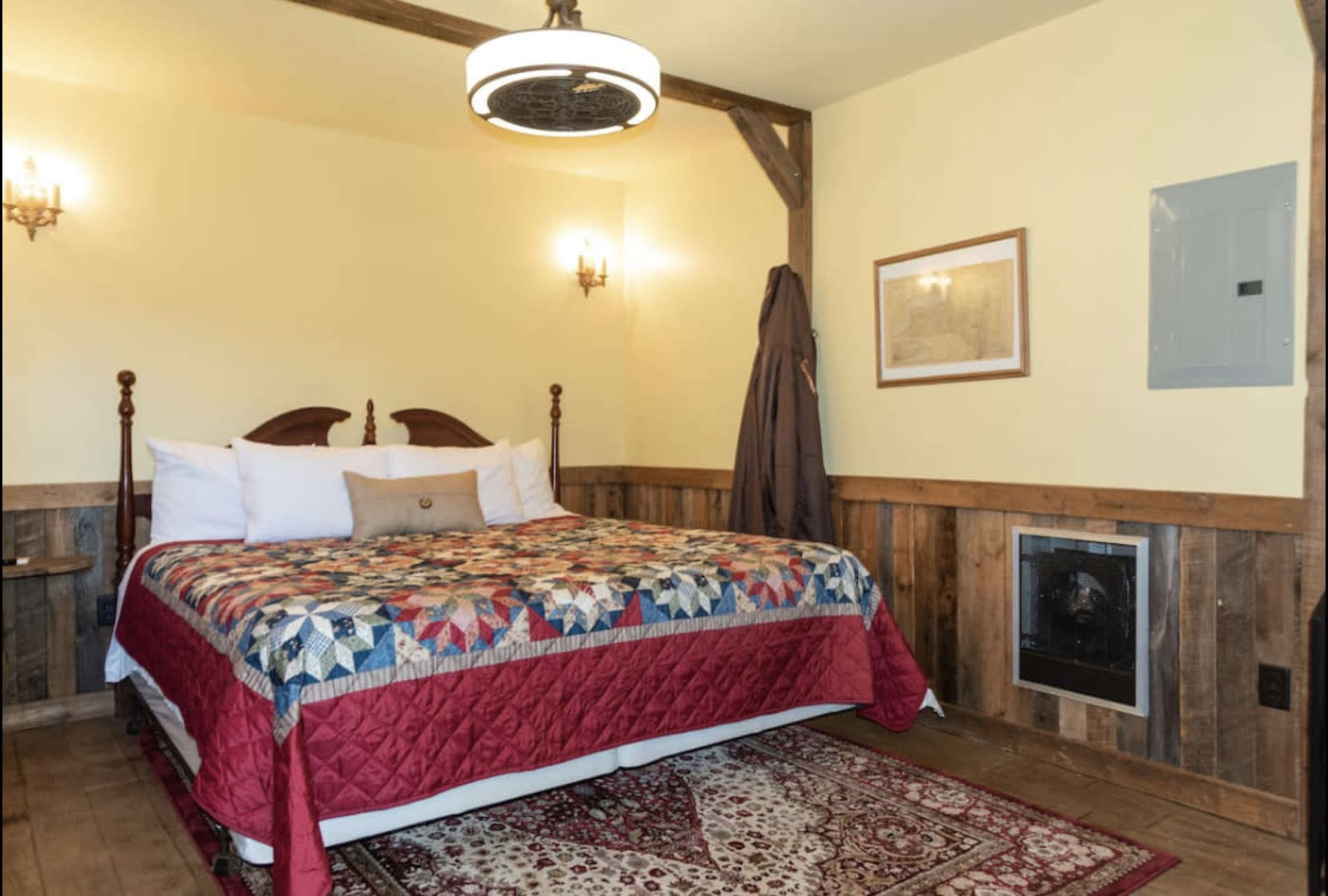A cozy bedroom featuring a large bed with a colorful quilt, wooden walls, a fireplace, and wall sconces for lighting.