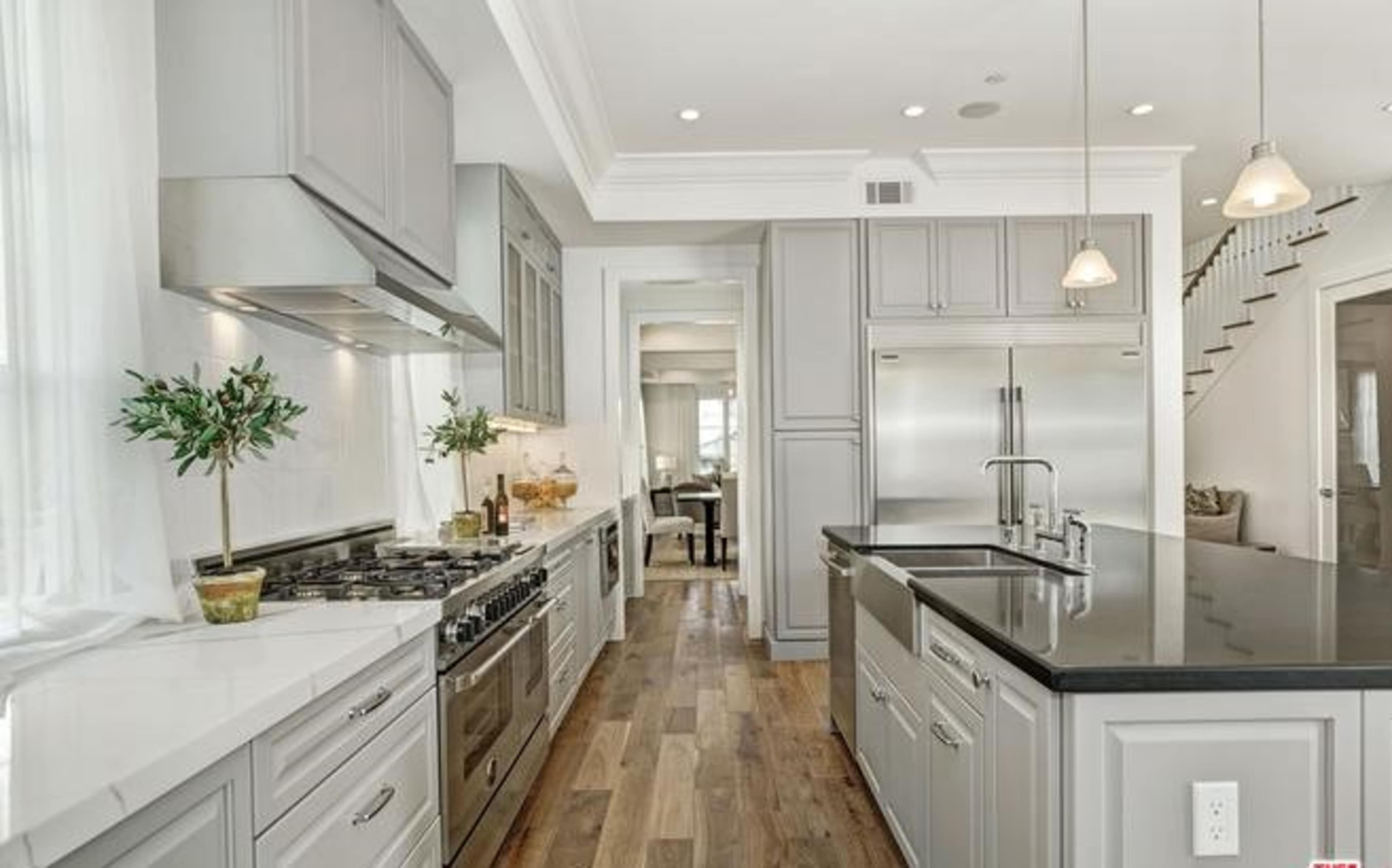 The image shows a modern kitchen featuring stainless steel appliances, gray cabinetry, and a dark countertop, with a wooden floor and a staircase in the background.