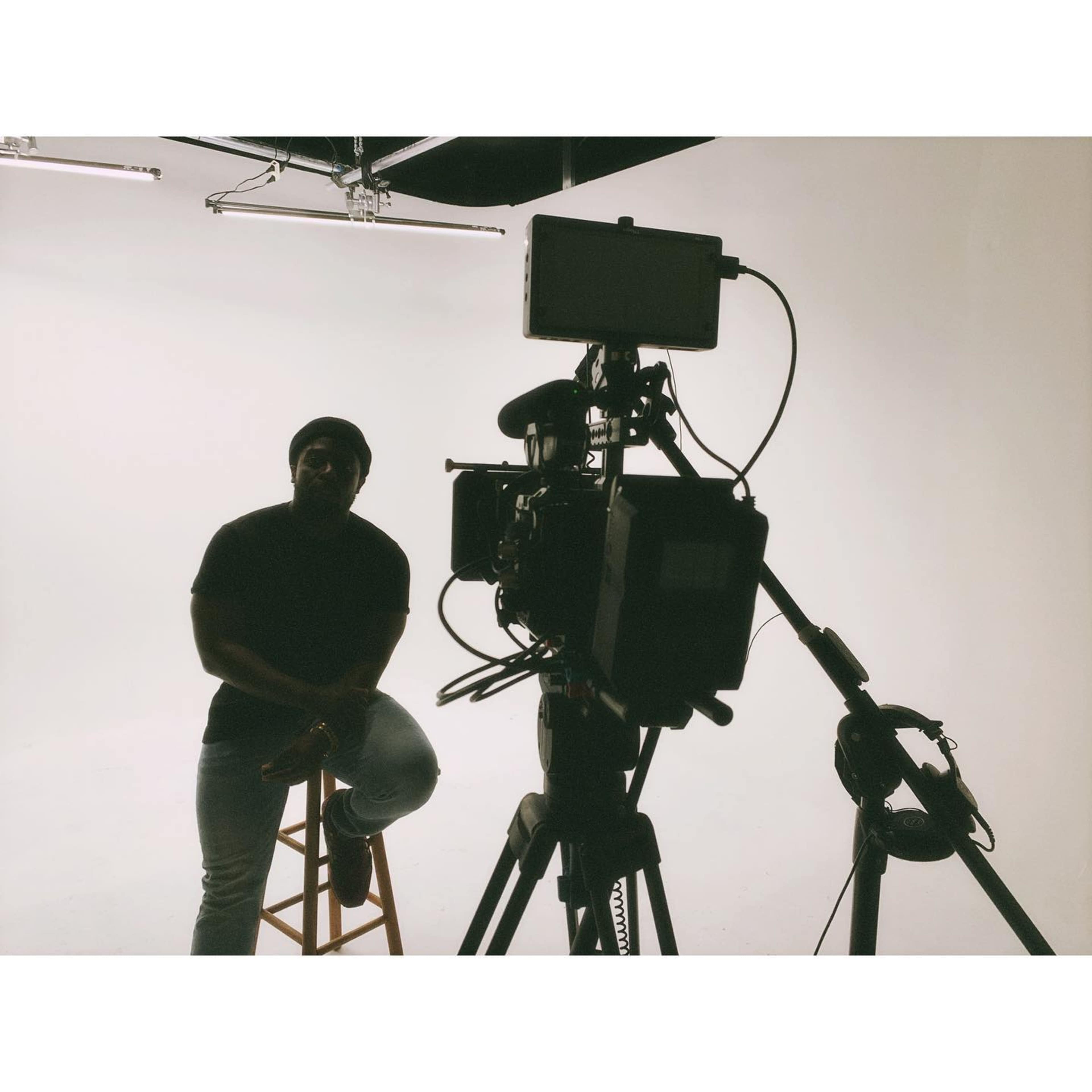 A person sits on a stool in front of a camera set up in a plain studio with bright lighting.