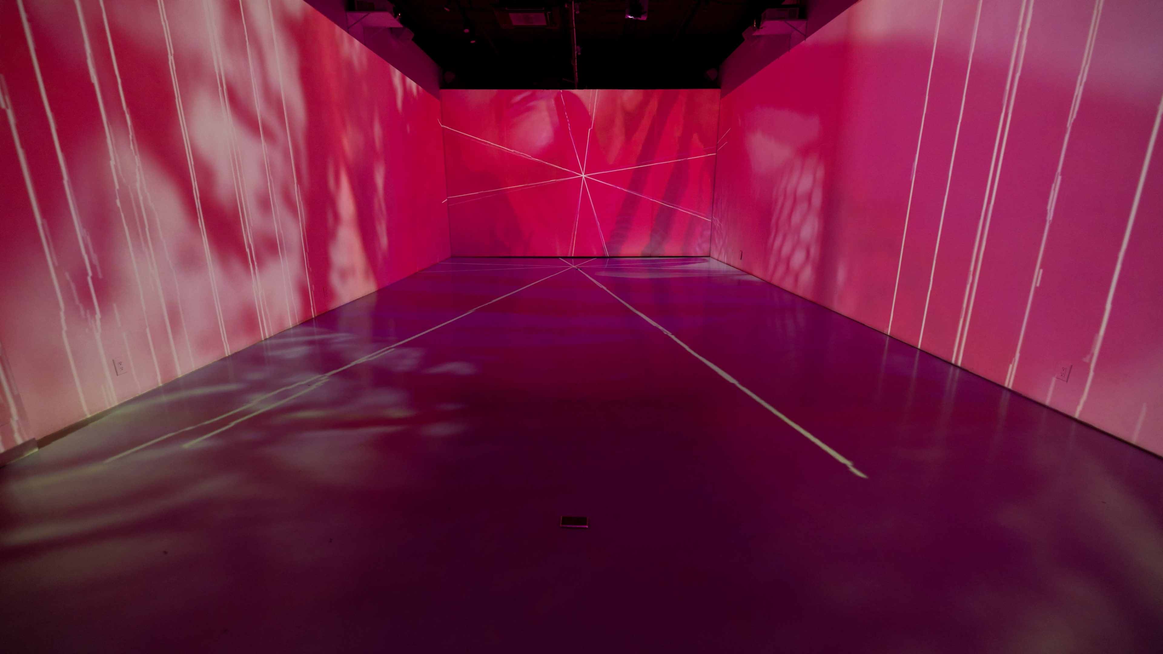 An empty room with pink-lit walls featuring projected shadows and intersecting lines on the floor.