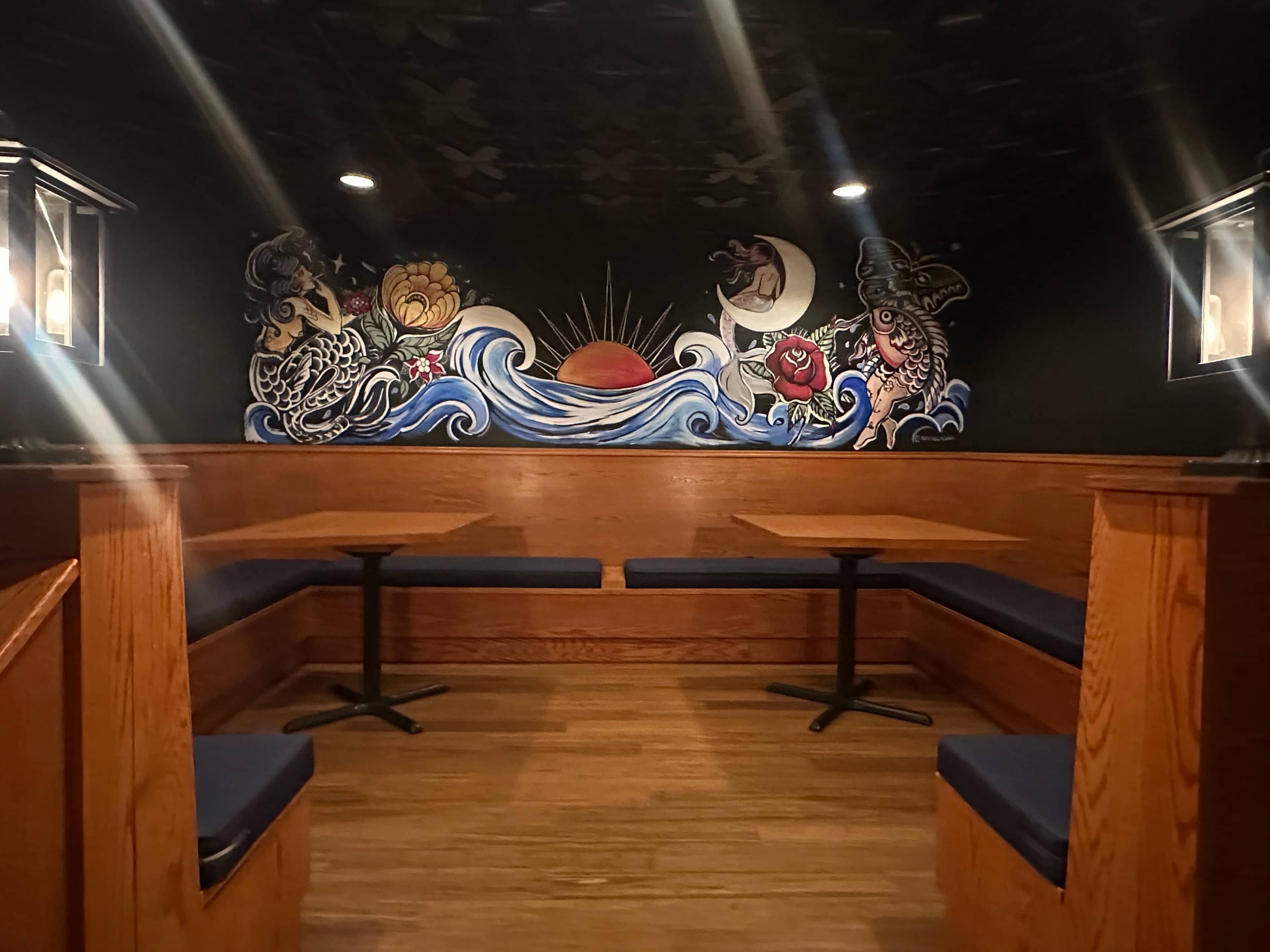 A cozy seating area in a dimly lit room featuring wooden booths and a colorful mural of waves and celestial elements on the wall.