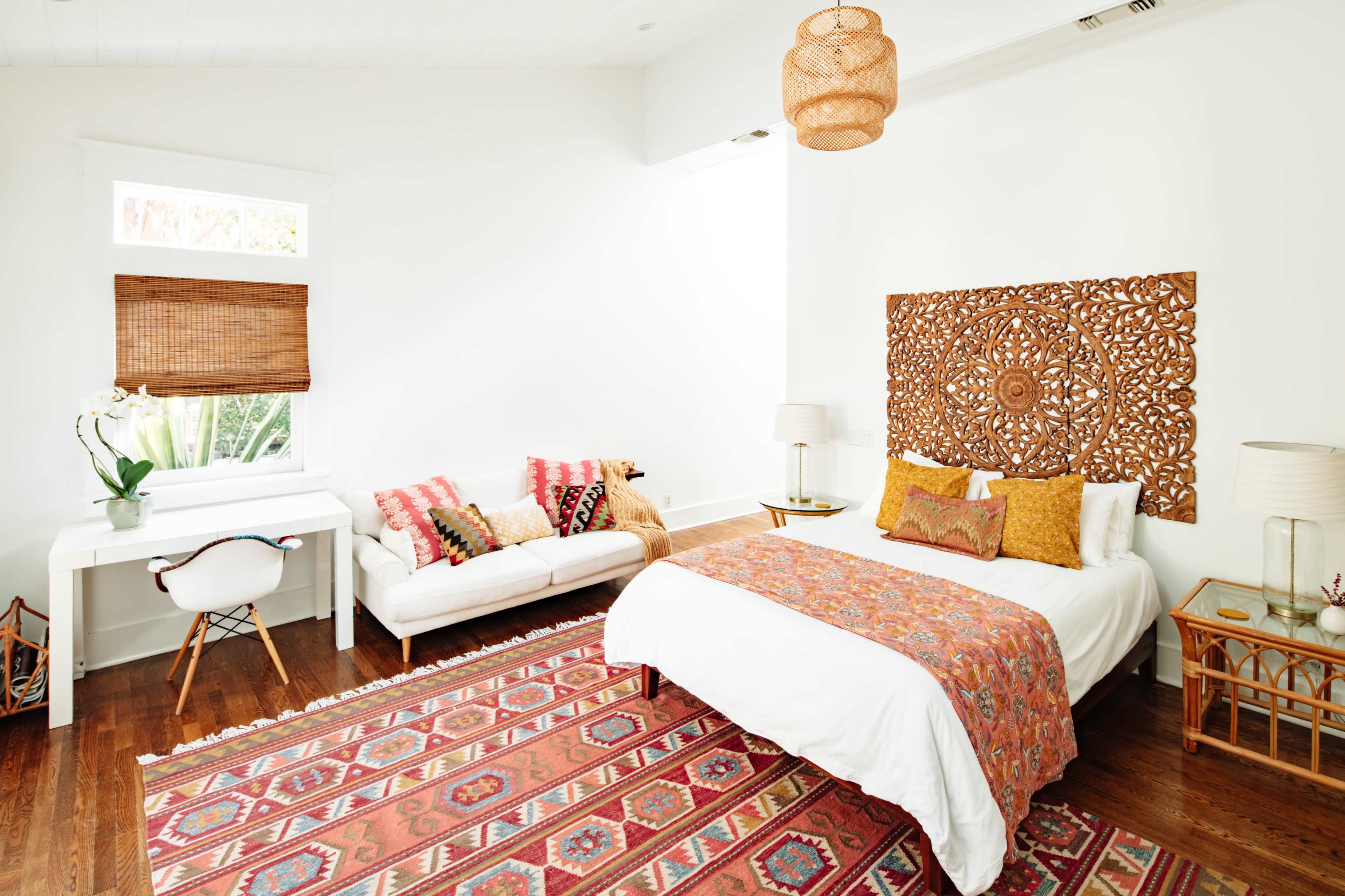 A bright, modern bedroom featuring a white bed with patterned bedding, a decorative wooden headboard, a cozy sofa with colorful cushions, a small desk, and a large area rug.
