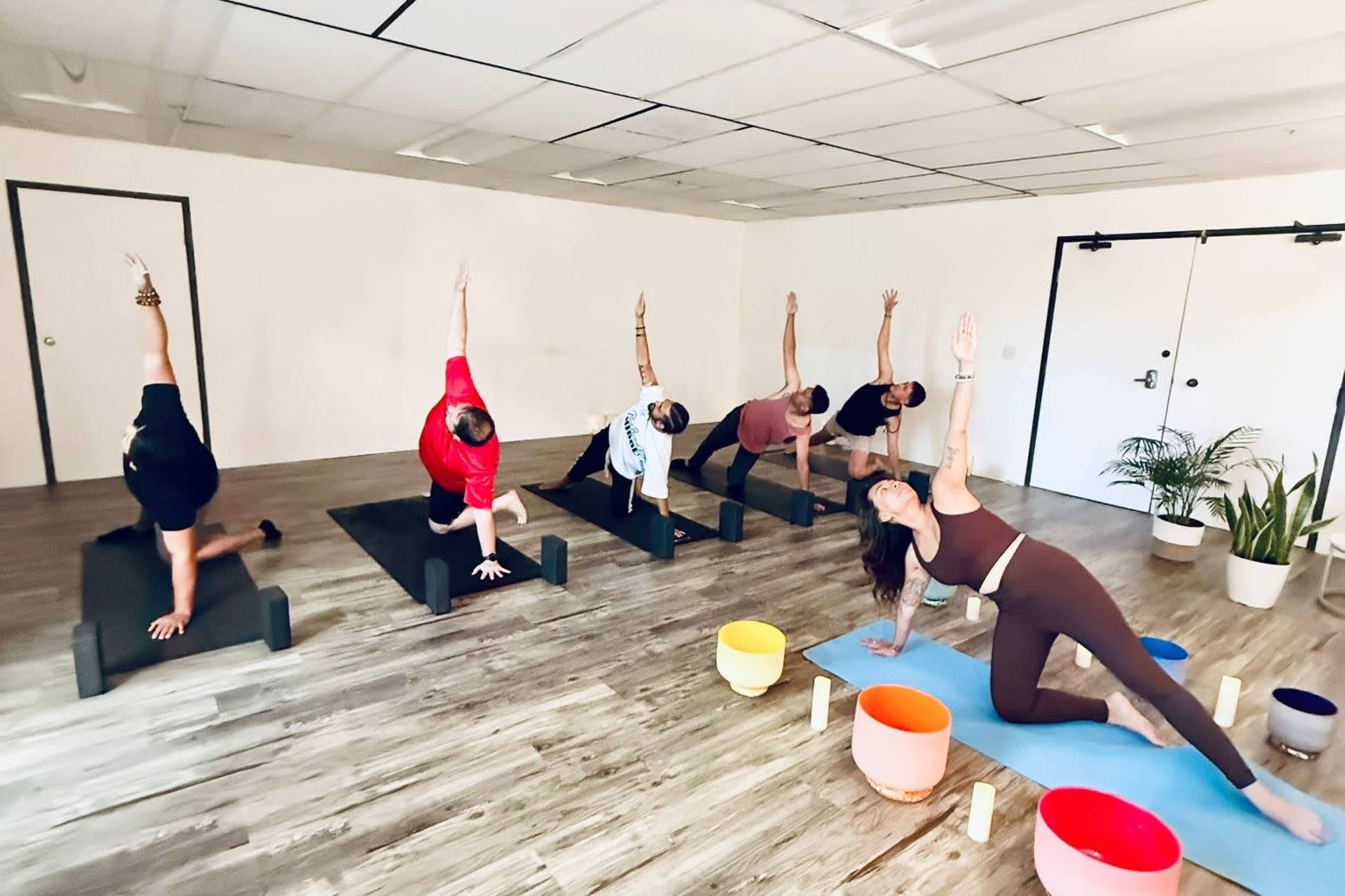 A group of individuals is performing yoga poses on mats in a bright studio with wooden floors and various props.