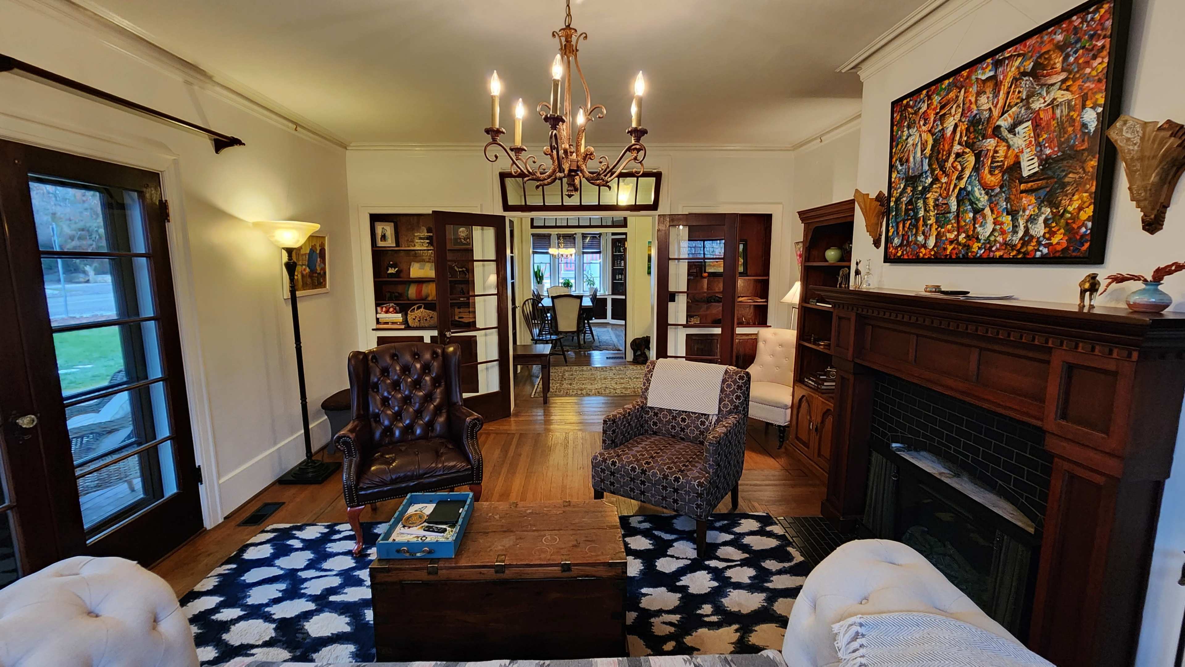 A cozy living room with a chandelier, a brown leather armchair, a patterned chair, a wooden coffee table, and a fireplace, all arranged around a decorative area rug.