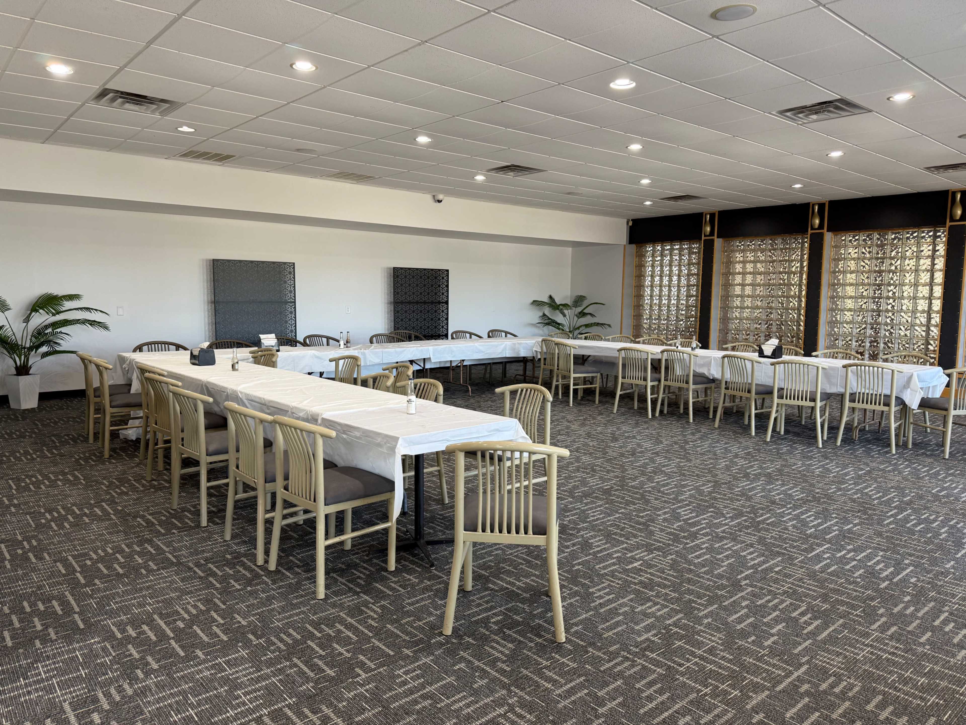 The image shows a spacious dining room with several long tables arranged in rows, featuring chairs and a carpeted floor.