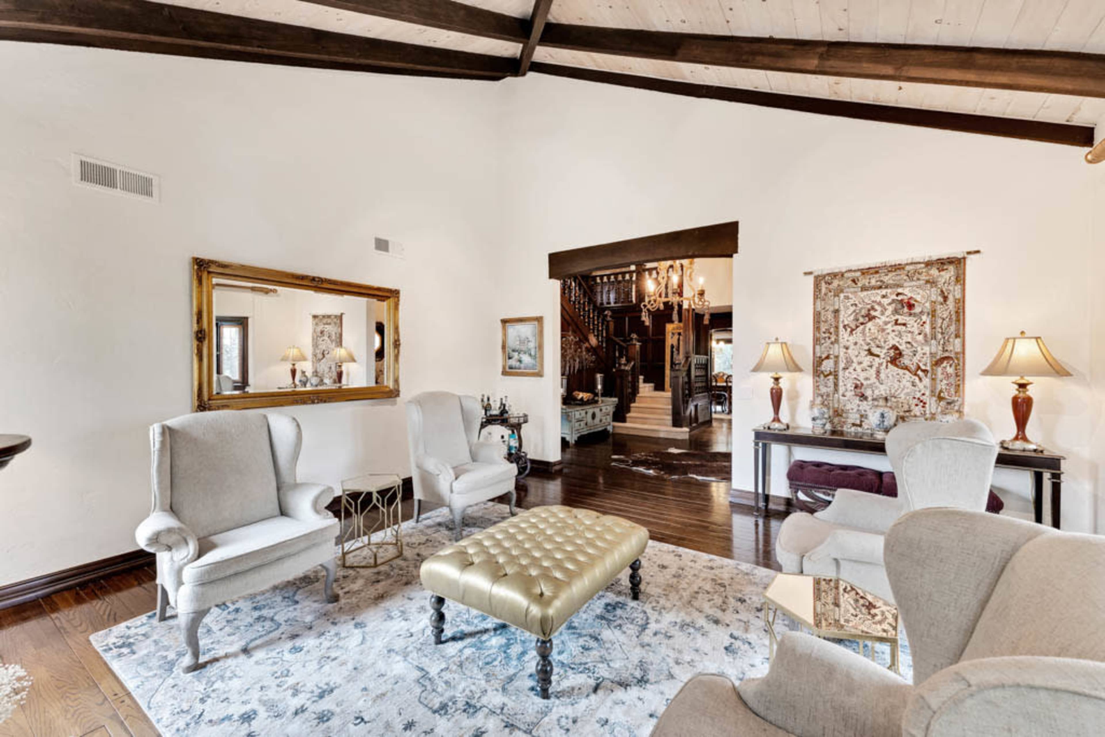 A spacious living room features cream-colored armchairs, a gold-tufted ottoman, and an ornate mirror, with a staircase and chandelier visible in the background.