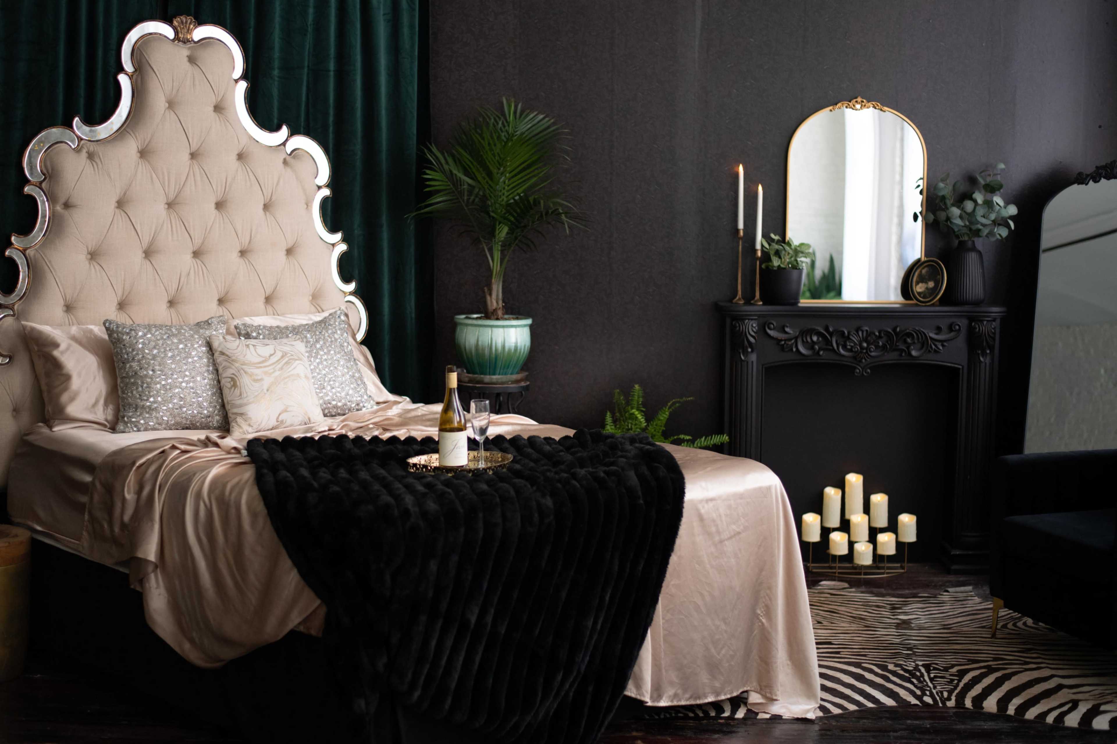 A luxurious bedroom features a large tufted headboard, a silk bedspread, decorative pillows, a mirrored nightstand, and a black faux fur throw, with candles and a plant arranged nearby.