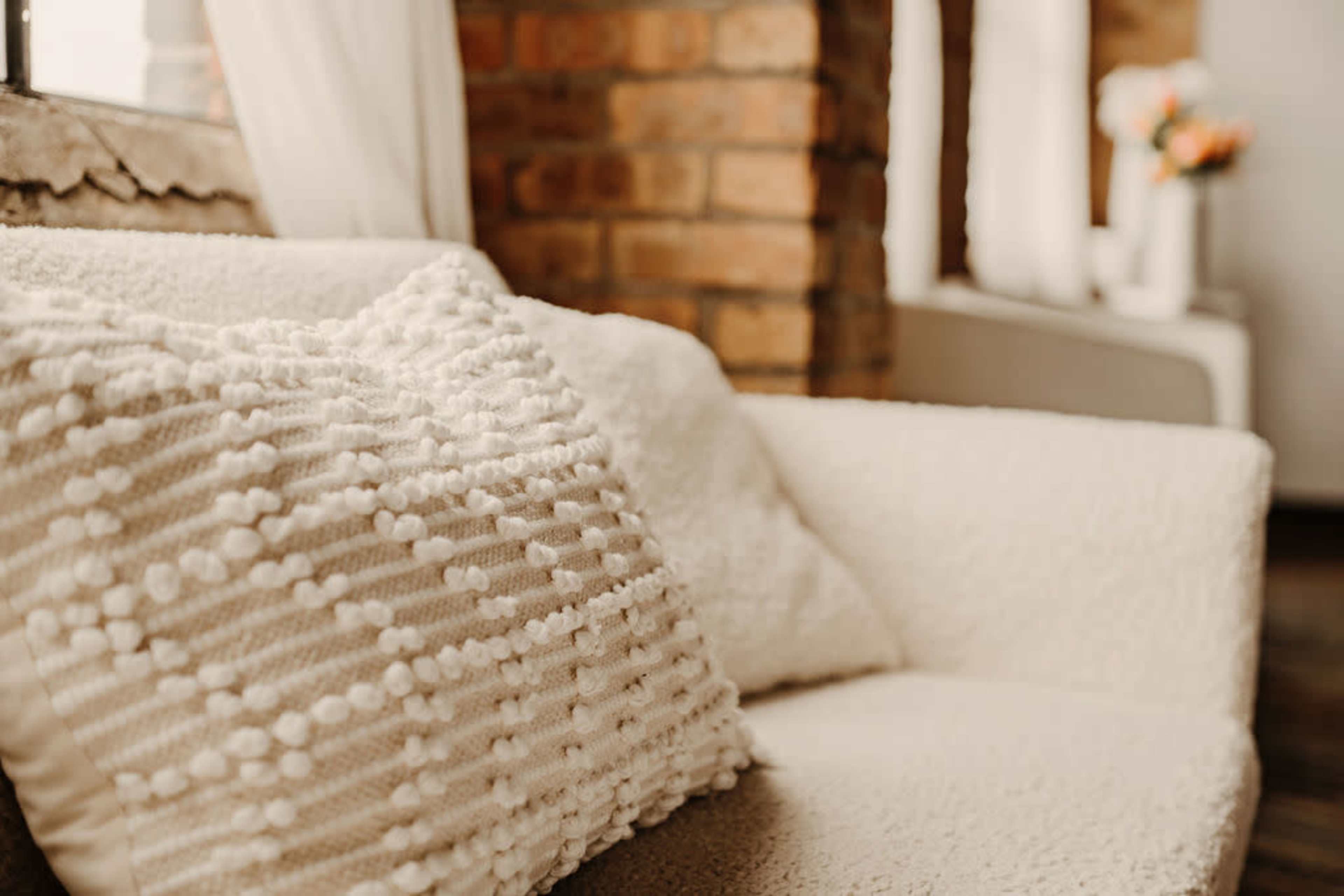 A cream-colored couch with a textured pillow is positioned near a brick wall and a window.