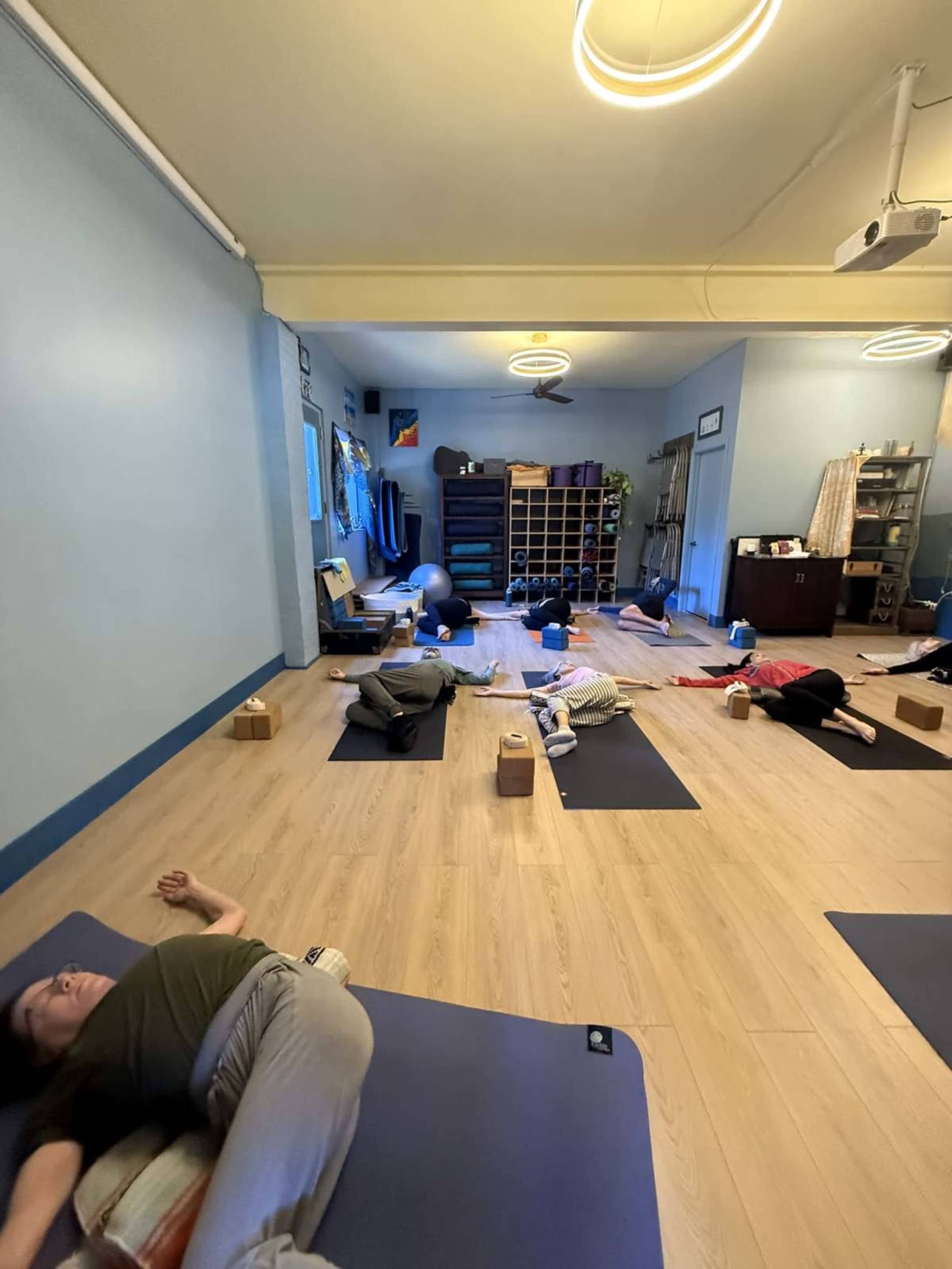 The image shows a yoga studio with several individuals practicing yoga on mats, some using blocks as props, in a calm and spacious environment.