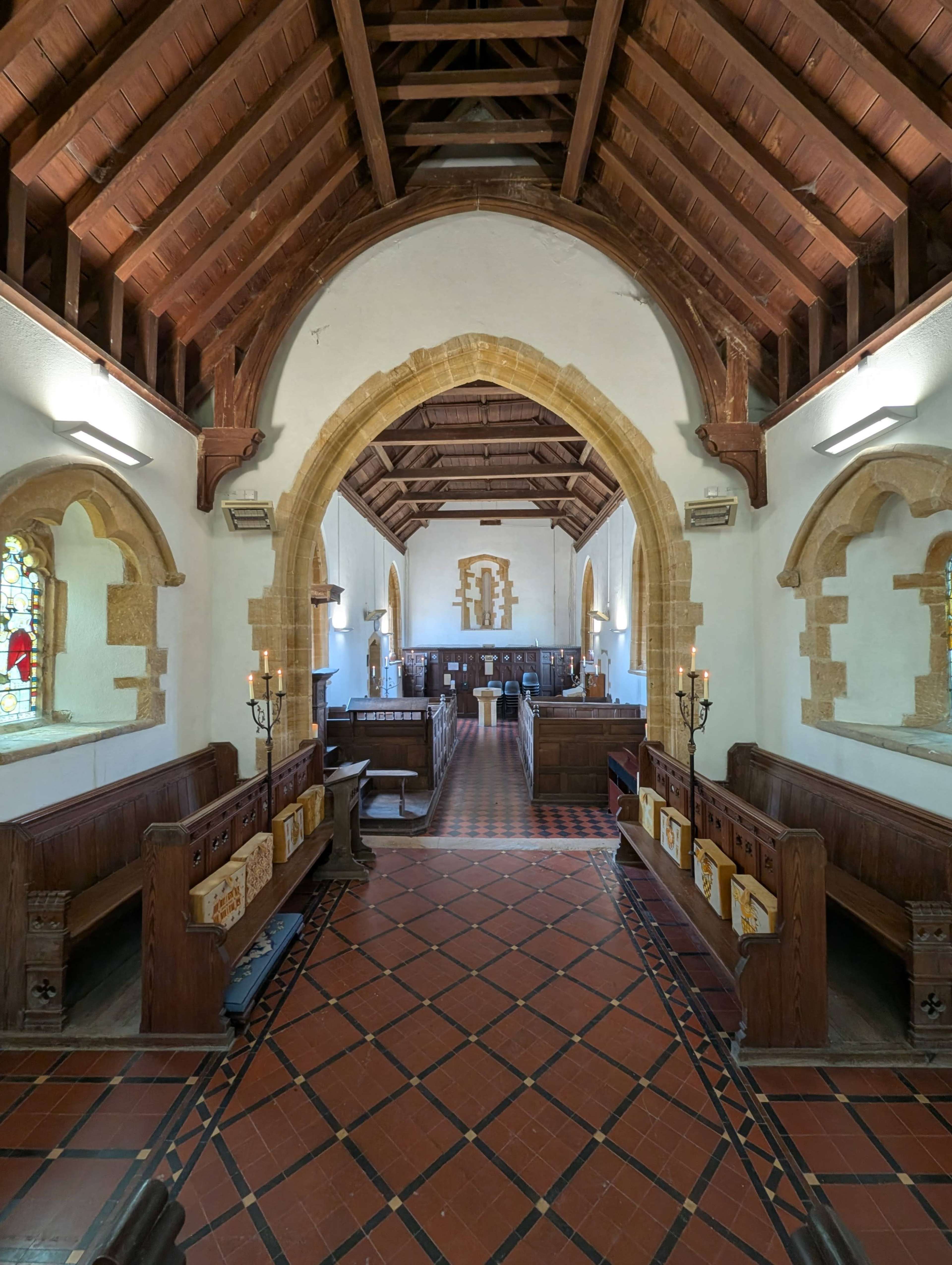 The image shows the interior of a church featuring wooden beams, arched doorways, and rows of wooden pews along with a tiled floor.