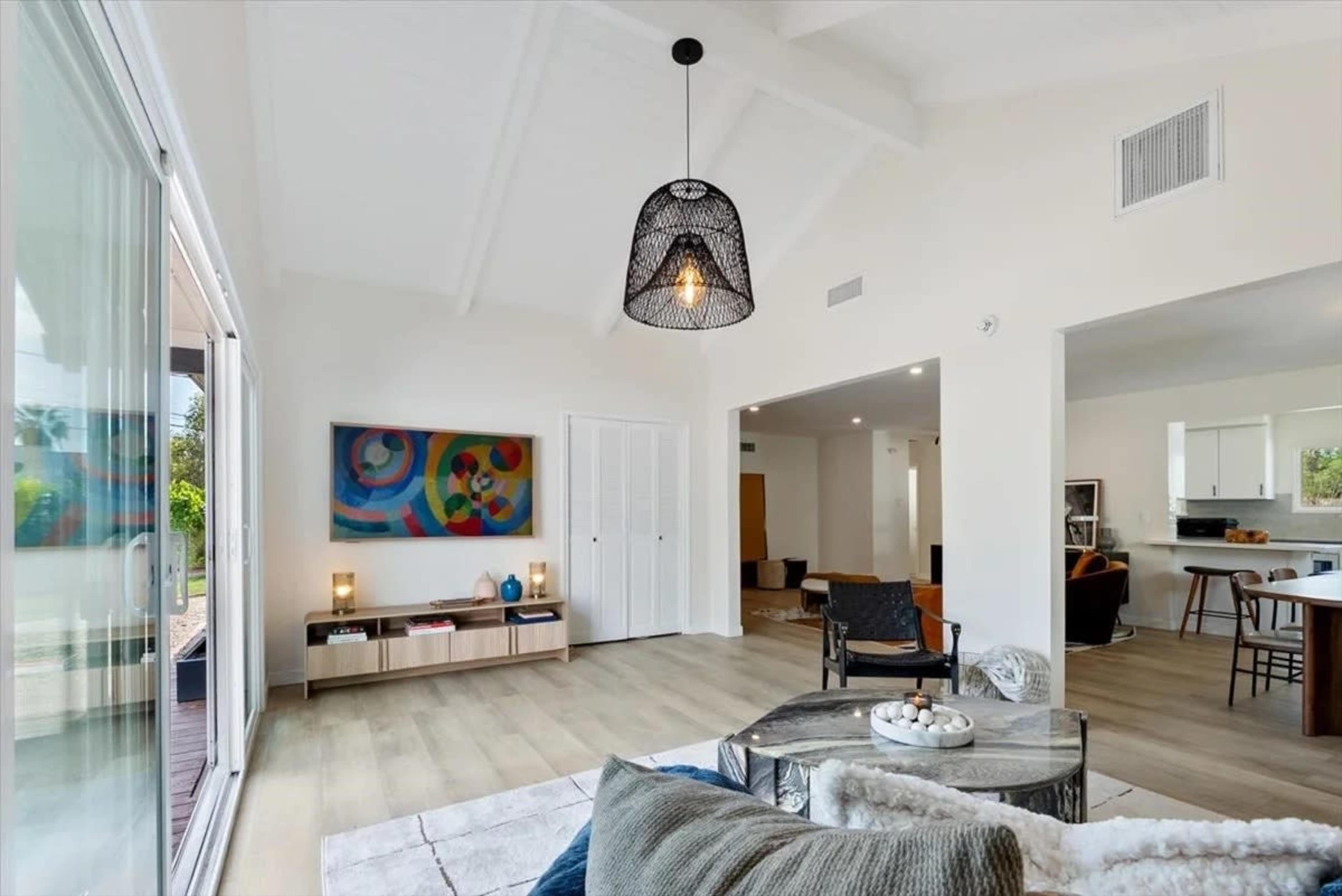 The image shows a modern interior living space with a high ceiling, a large wall-mounted artwork, and an open layout leading to a kitchen area.