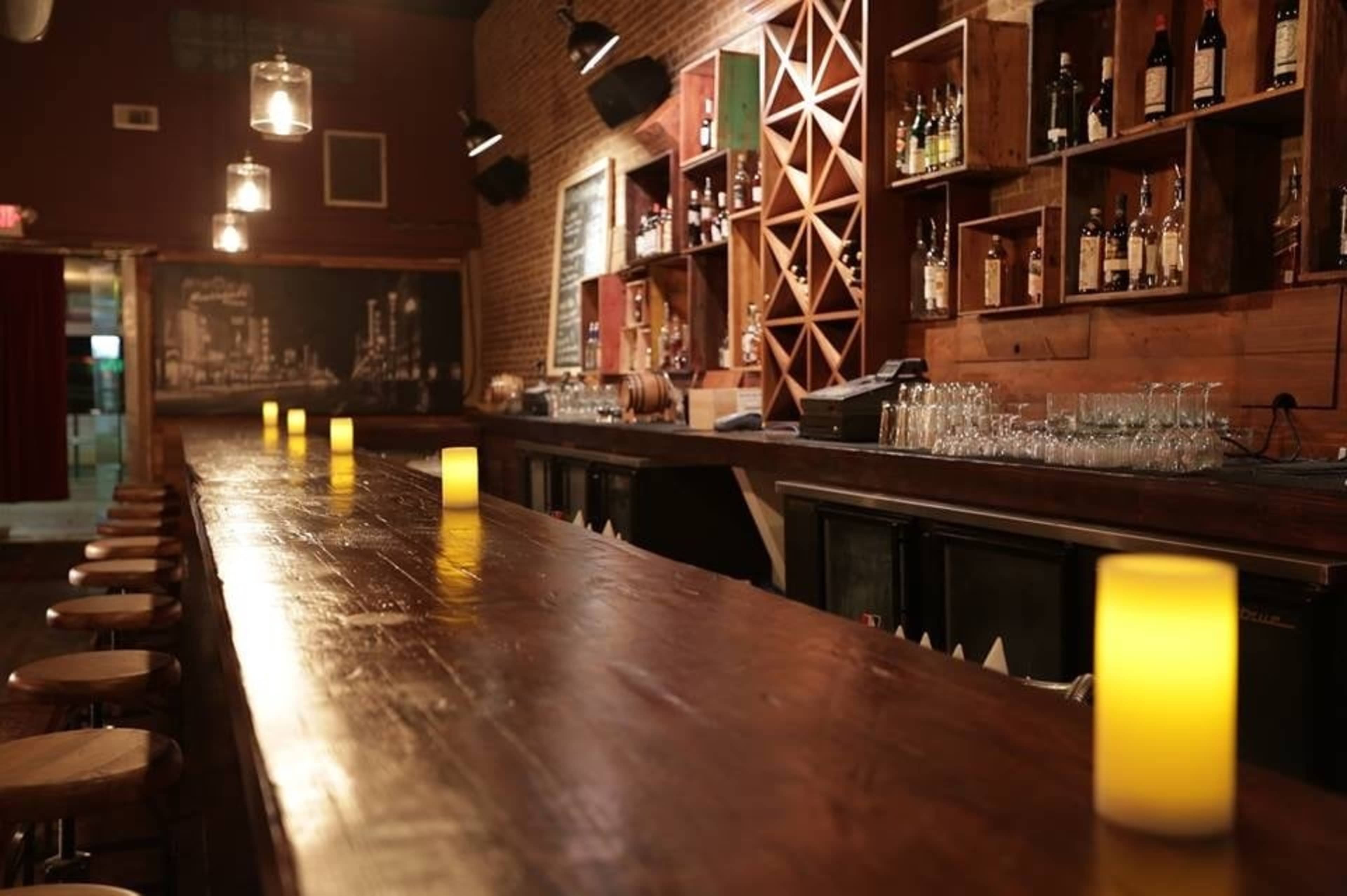 A wooden bar with a long countertop, illuminated by glowing candles, surrounded by shelves filled with various bottles.