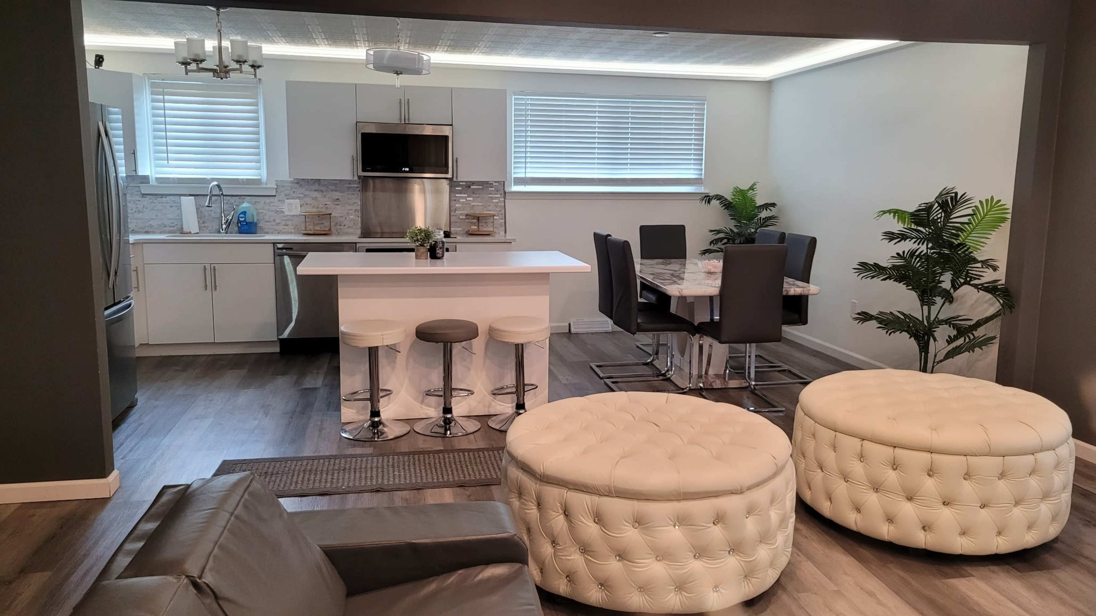 A modern kitchen and dining area feature sleek cabinetry, a marble island, and two tufted ottomans in a cozy seating arrangement.