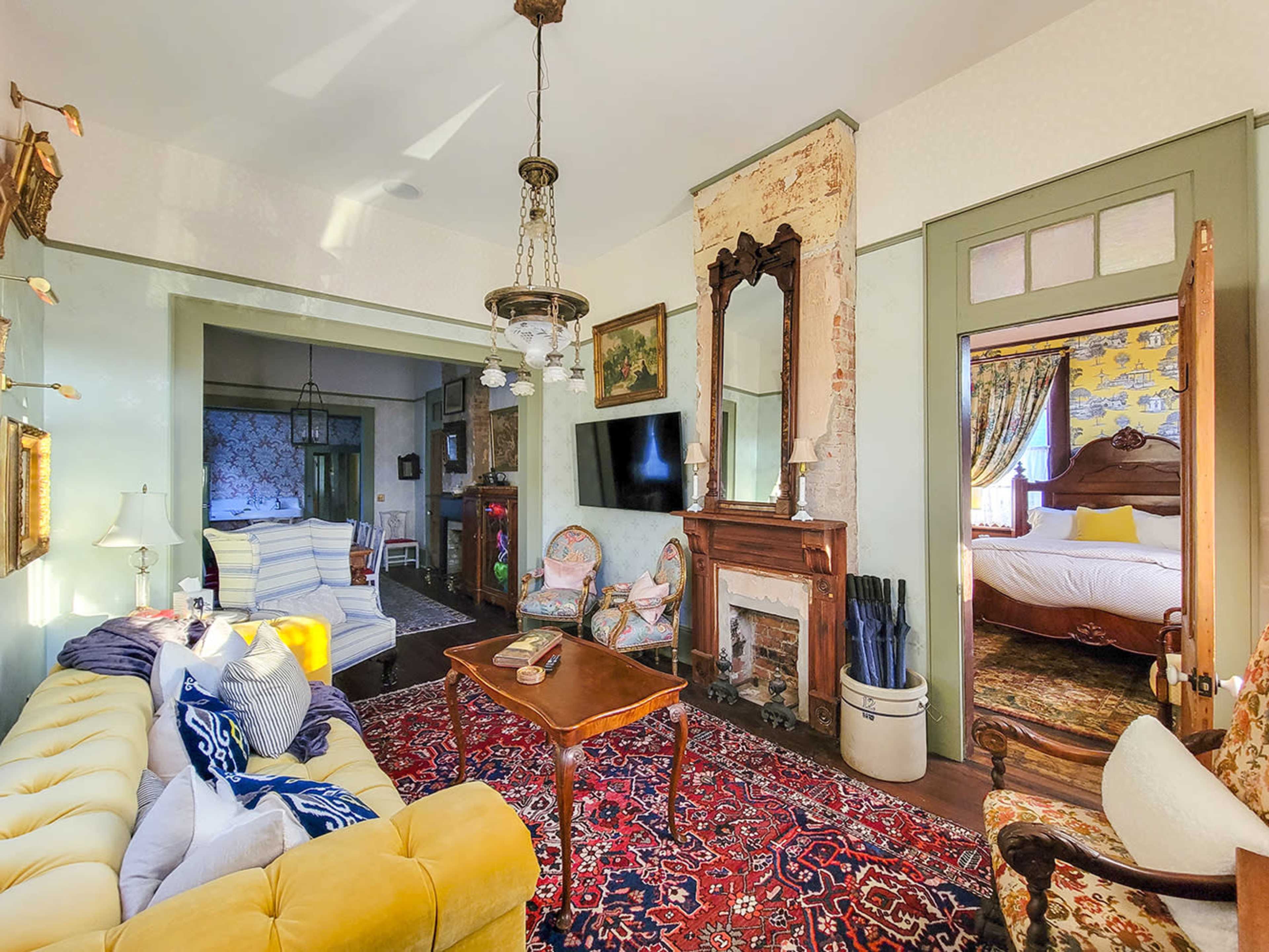 A cozy living room with a yellow sofa, a wooden coffee table, and an ornate fireplace, leading to a room with a bed visible in the background.