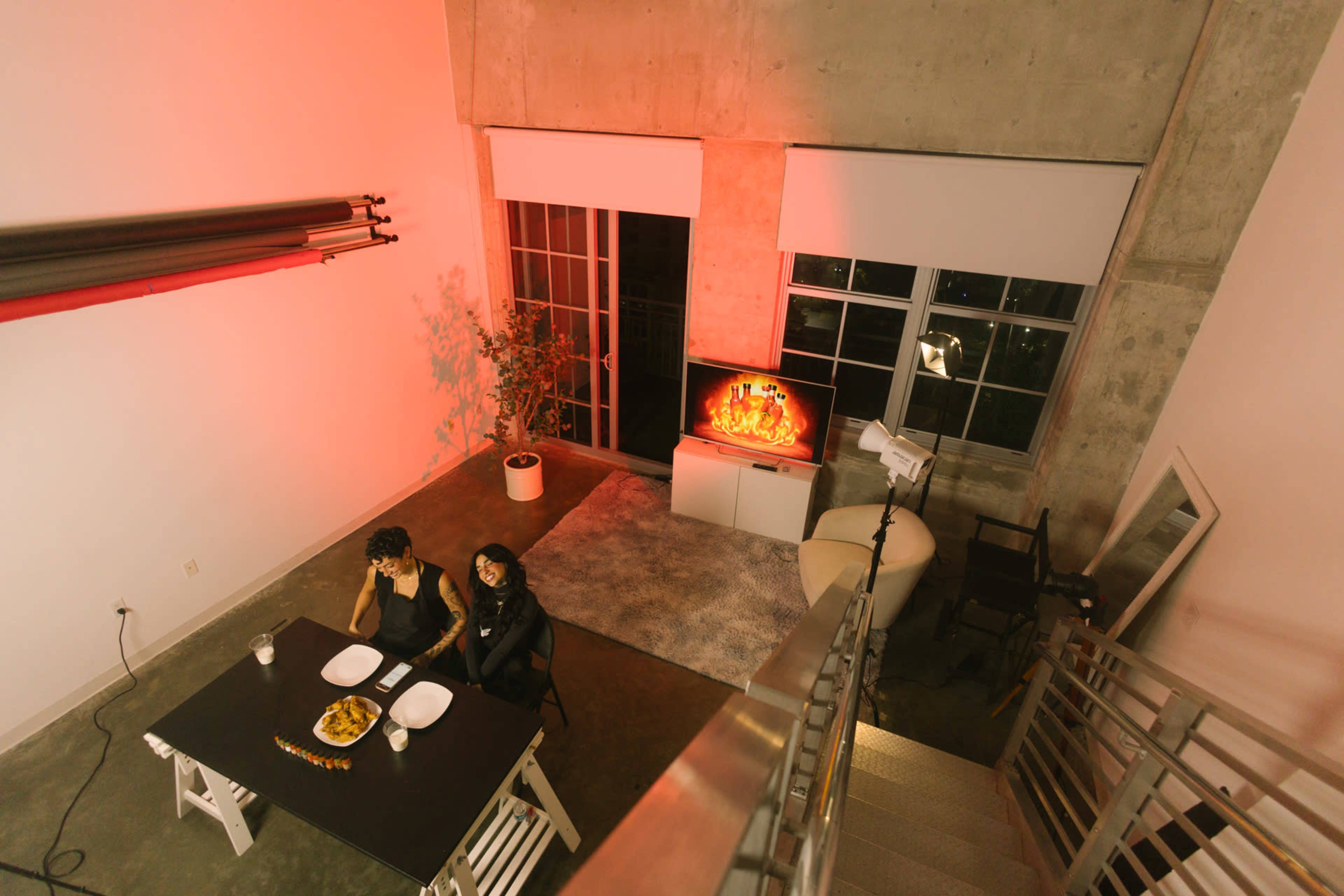 Two people sit at a black table with food in a modern, sparsely furnished room illuminated by soft red lighting and a television displaying a flickering image.