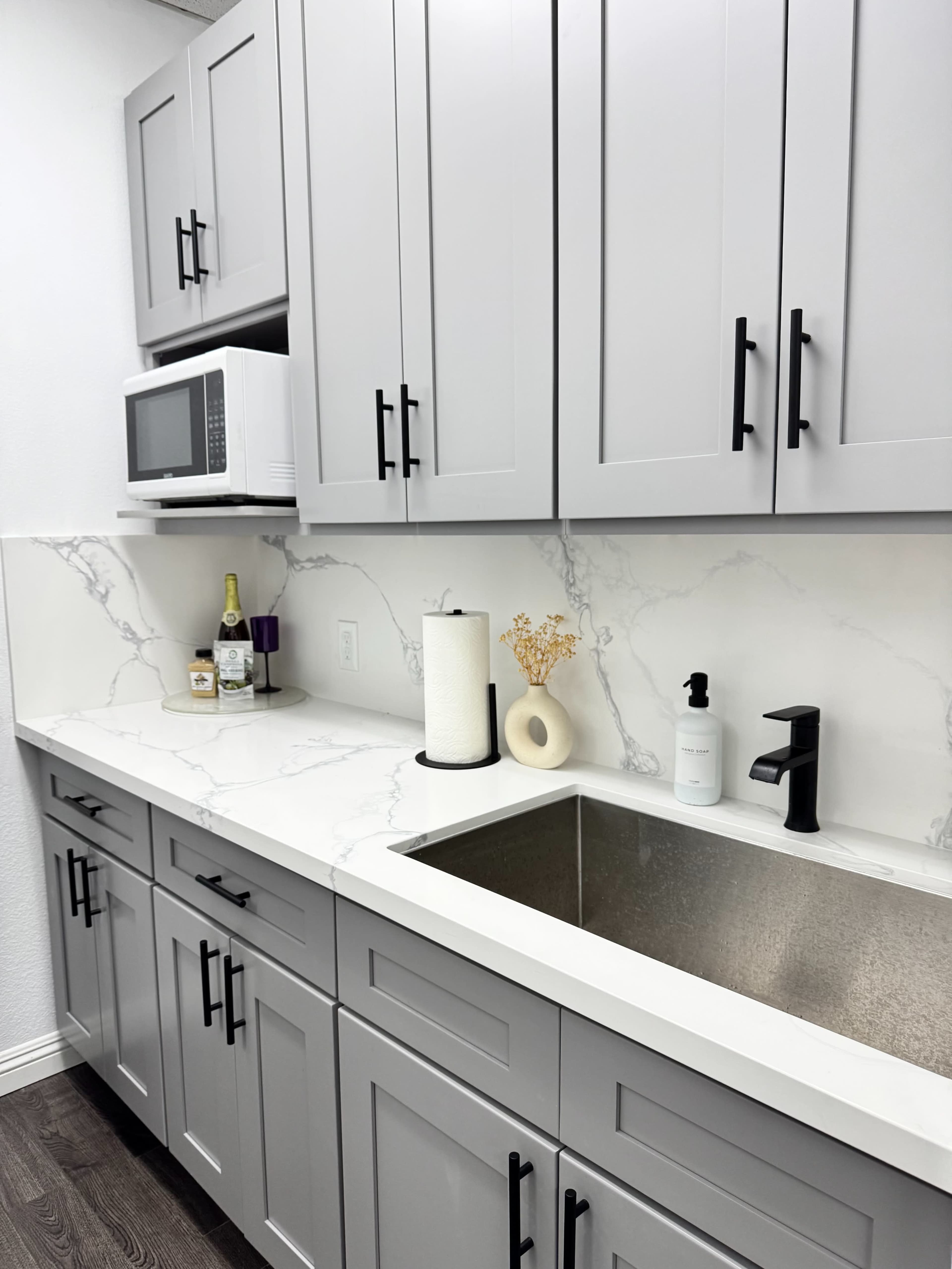 The image shows a modern kitchen with gray cabinets, a stainless steel sink, and a white countertop featuring a marble design, along with a microwave and decorative items on the counter.