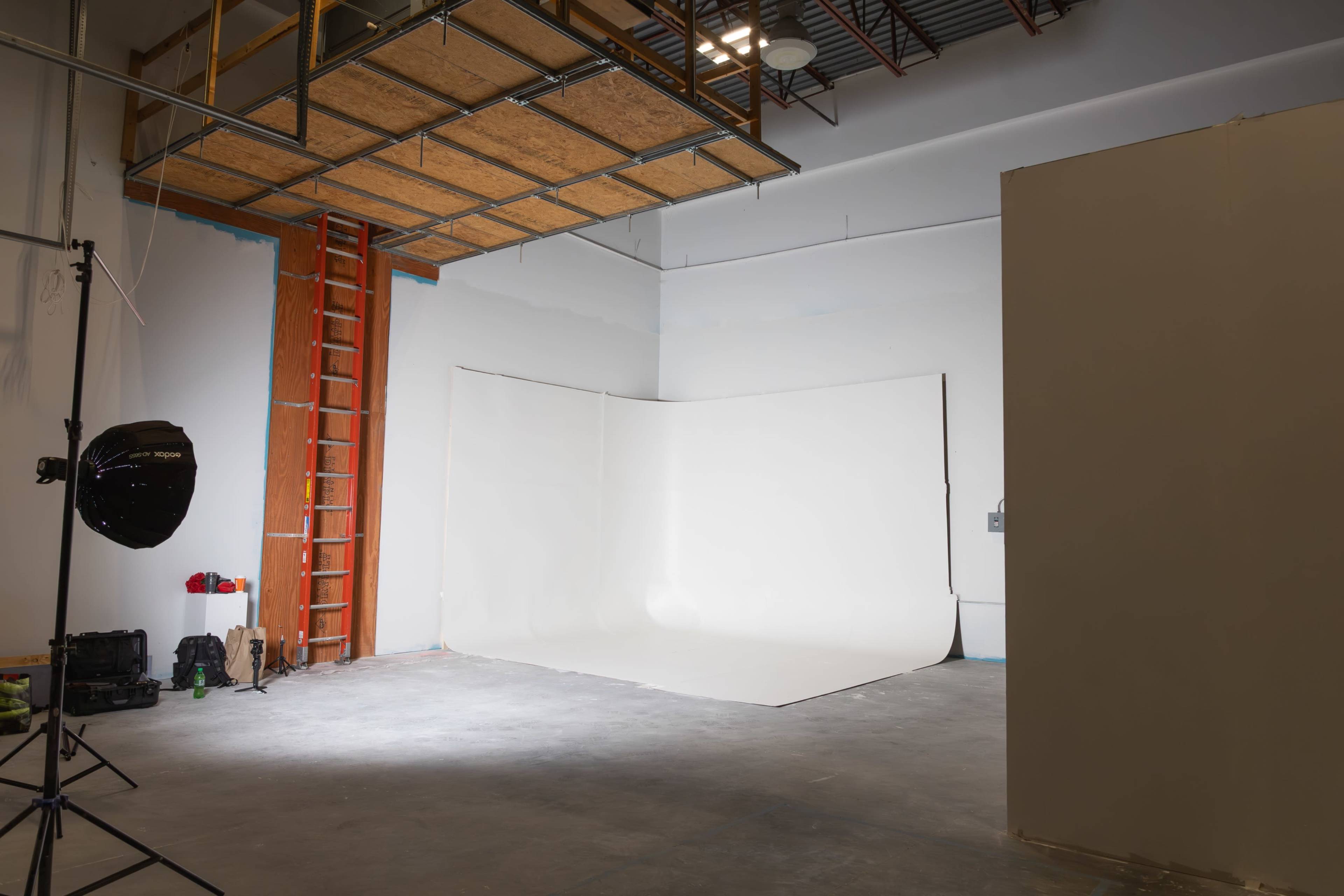 The image shows a spacious photography studio with a white backdrop, a ladder, and lighting equipment.