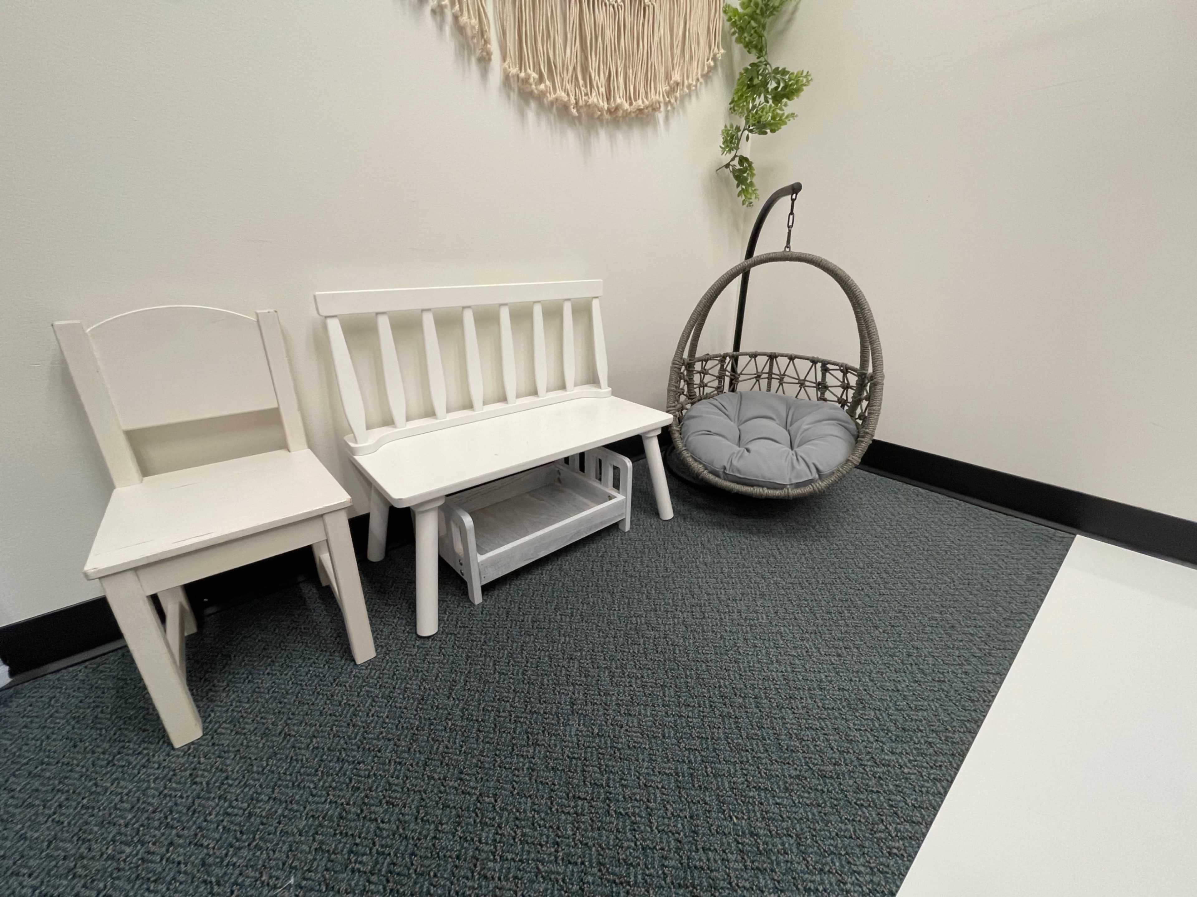 The image shows a small indoor area with a white bench, a single white chair, and a hanging, cushioned swing in a gray and green color scheme.